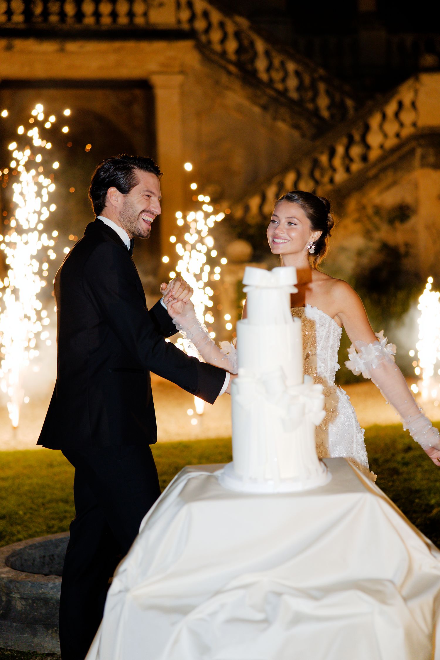 Joyful newlyweds celebrate with sparklers next to their elegant white wedding cake at a historic venue.
