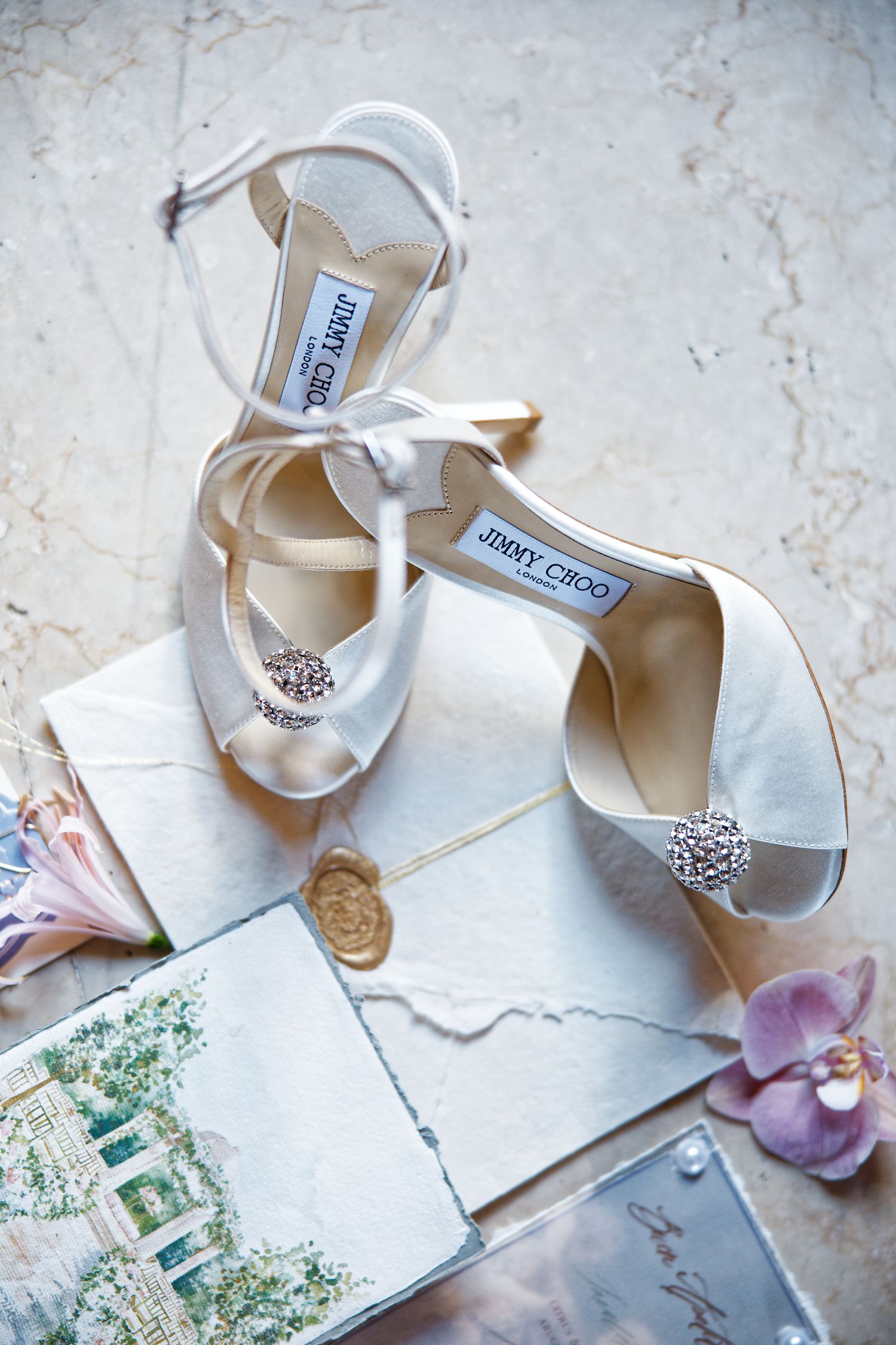 White Jimmy Choo bridal shoes with crystal embellishments displayed with vintage postcards and flower petals.