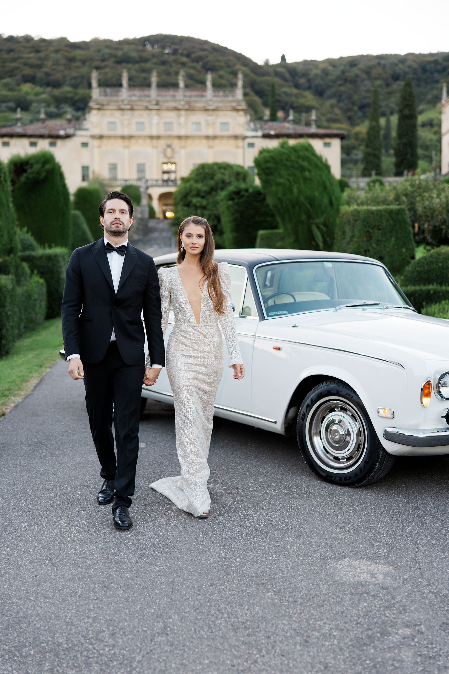 A couple in wedding attire pose in front of a classic white car and Italian villa.