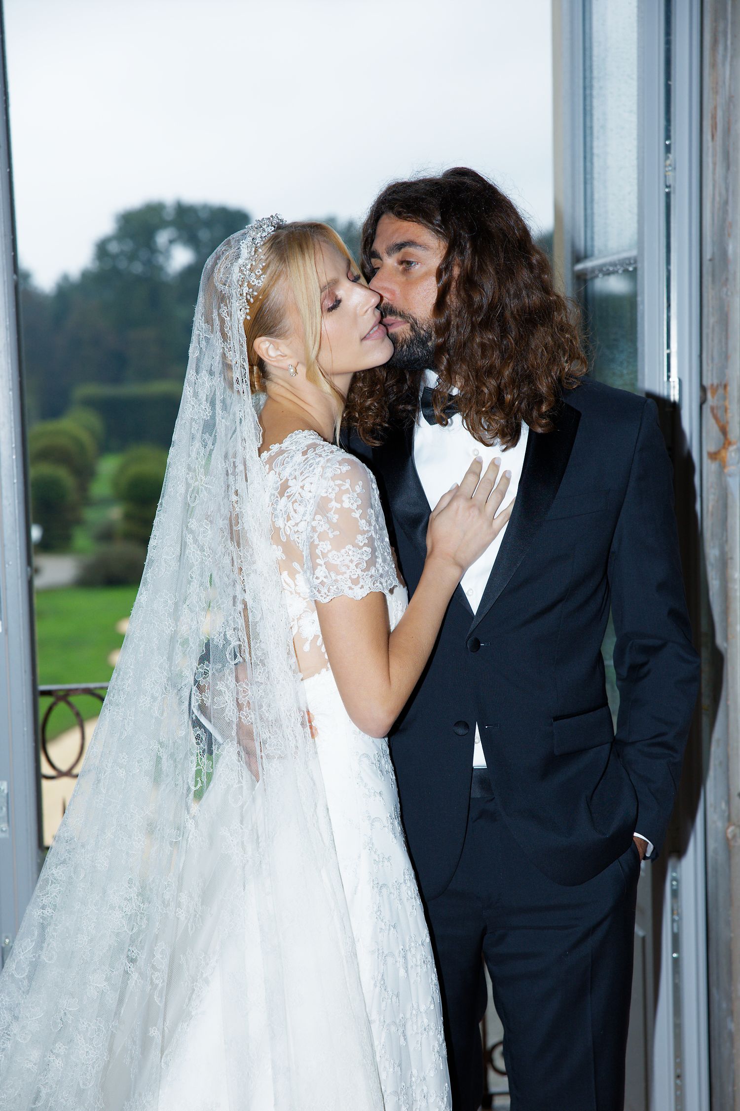 Romantic wedding moment captured by window with flowing white veil and formal attire.