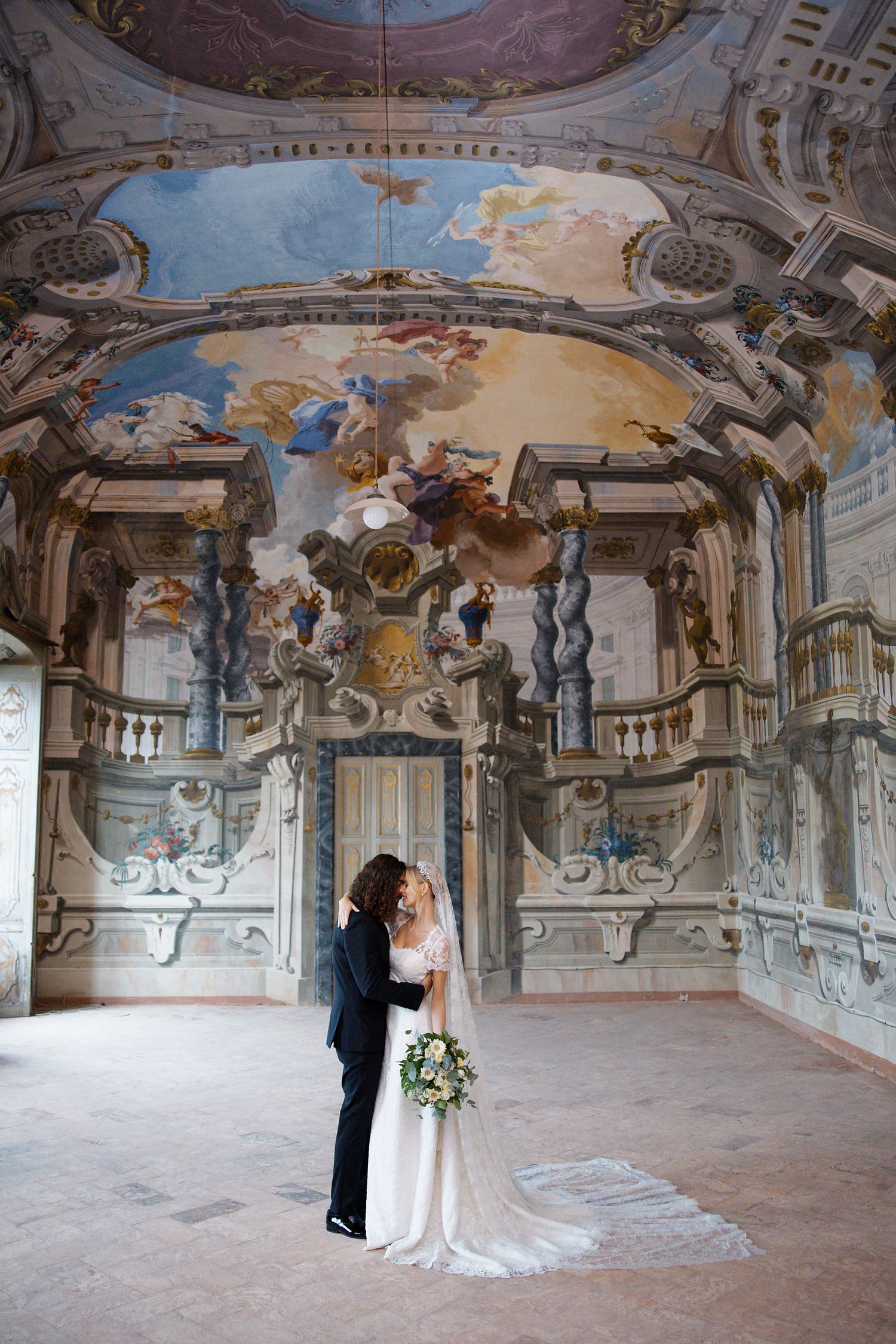 A wedding couple shares an intimate moment in an ornate baroque room with beautifully frescoed ceilings and marble columns.