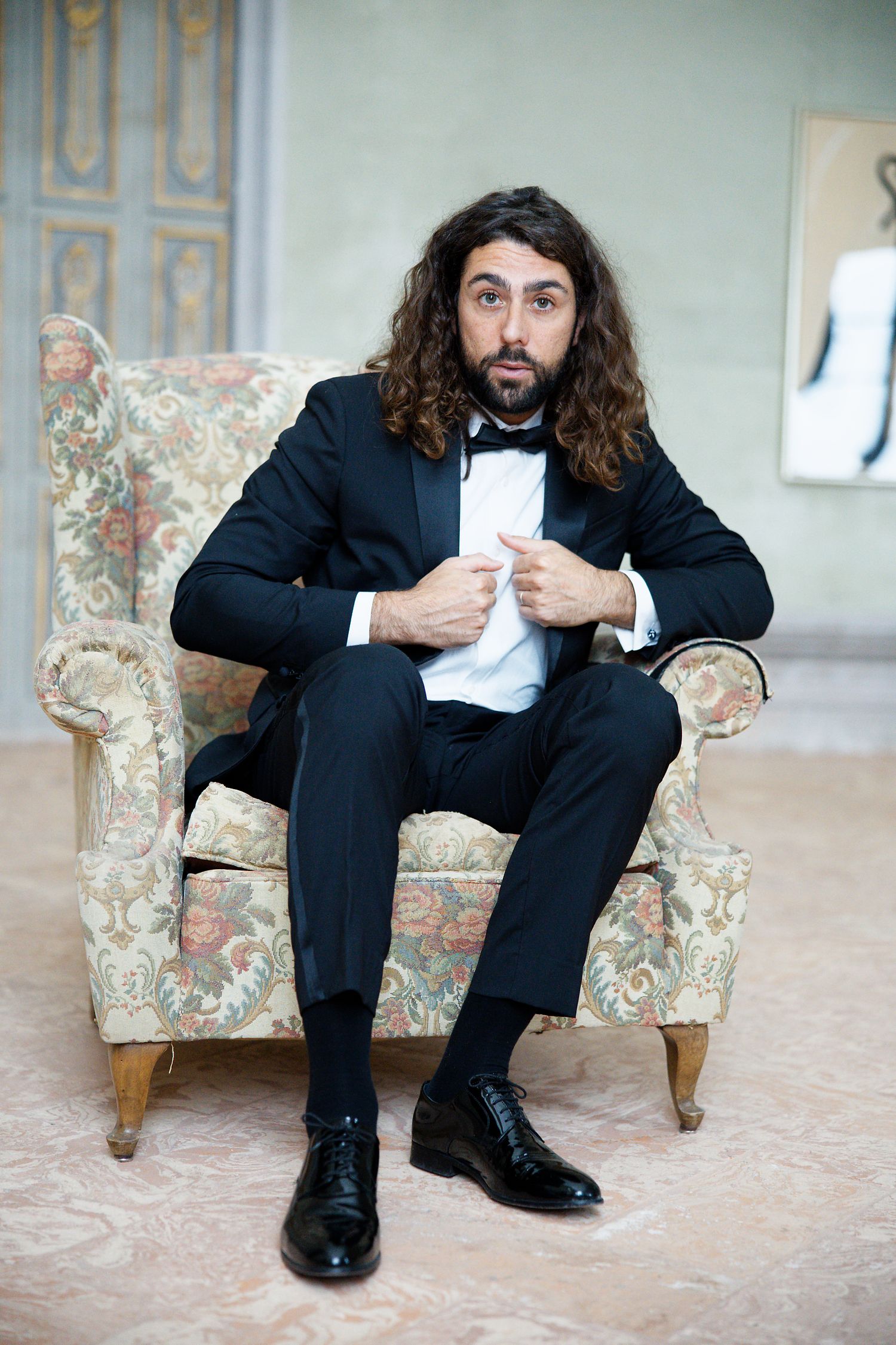 A bearded man in a black tuxedo sits on a floral upholstered armchair in an elegant setting.