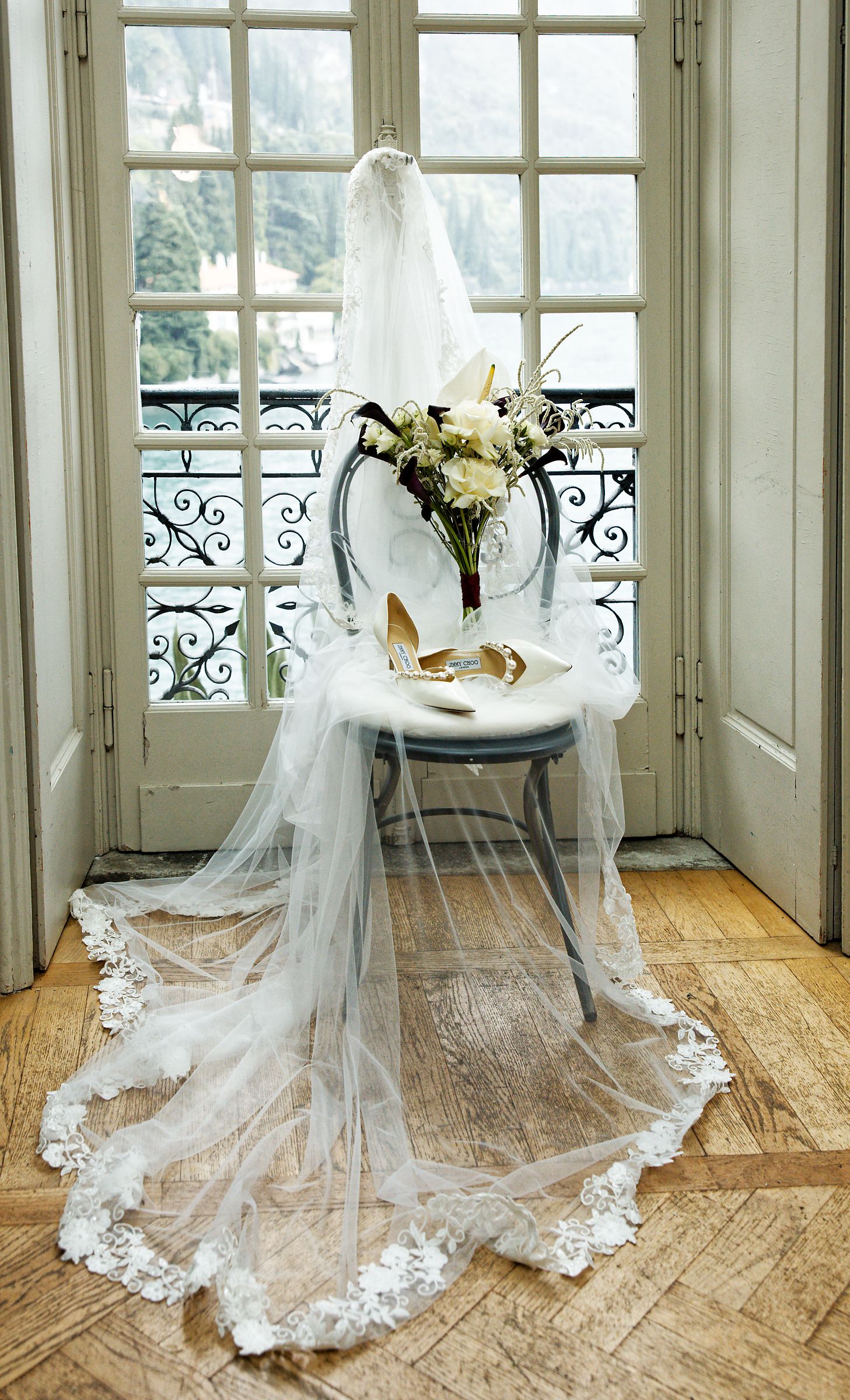 A white wedding veil and gold heels arranged on a chair by French doors with a bouquet of white flowers.
