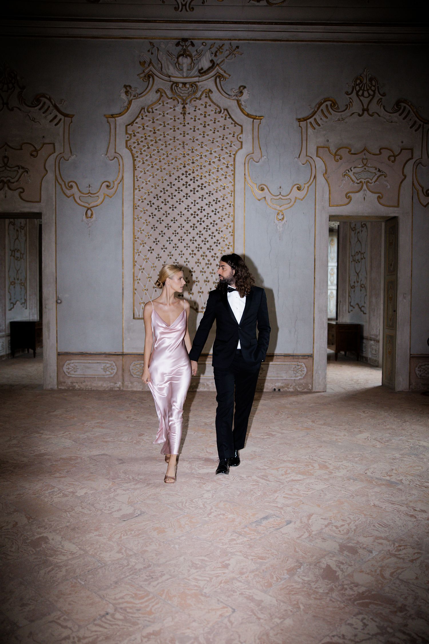 Elegant couple in pink silk dress and black tuxedo walking through ornate vintage ballroom.