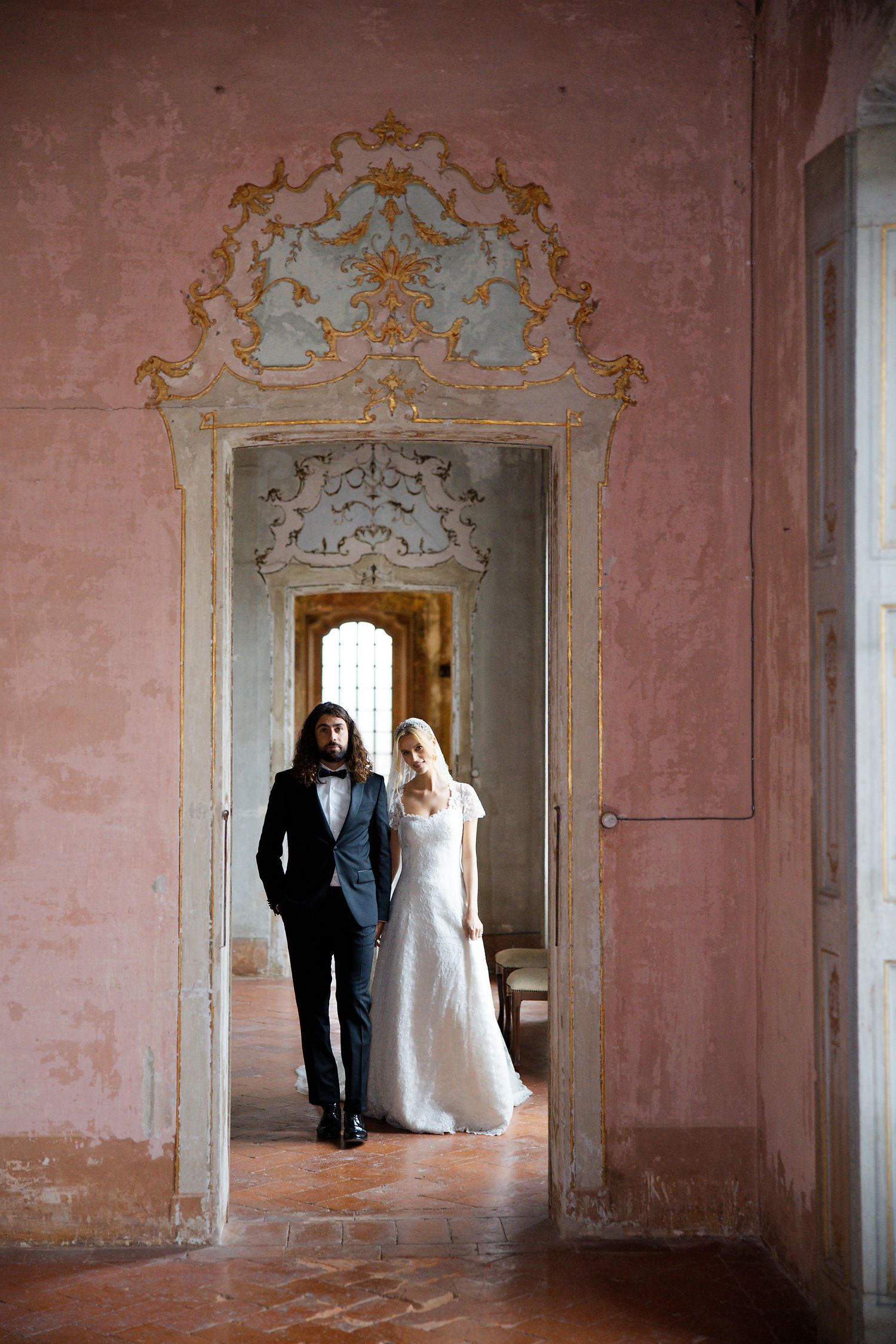 A couple standing in an ornate pink hallway with baroque architectural details and decorative moldings.