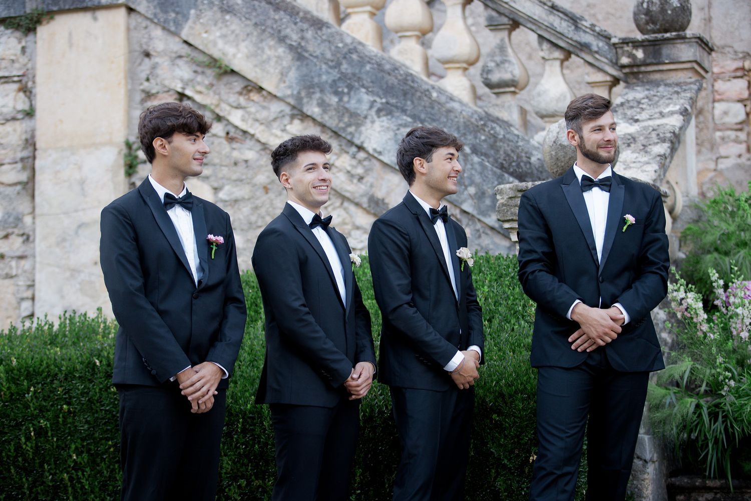 Groomsmen stand in formal line wearing black tuxedos against classic architecture.