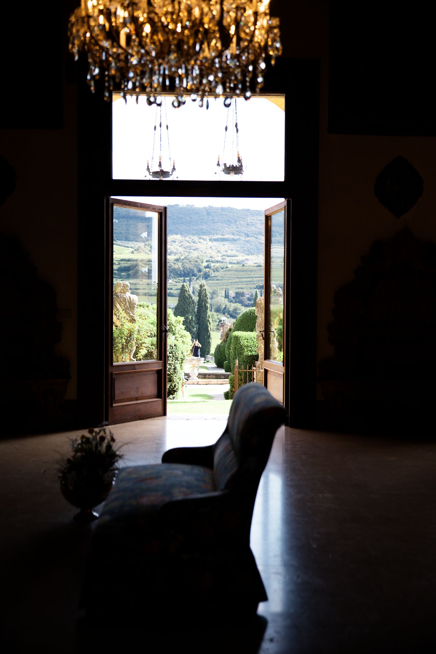 Elegant crystal chandelier hangs above window with scenic Tuscan countryside view.