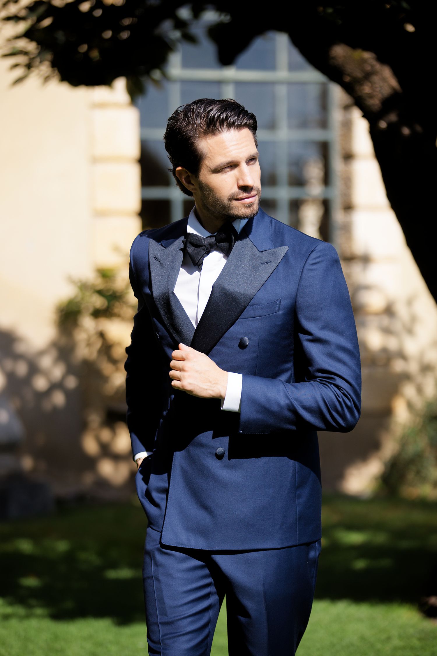 Stylish groom in navy blue double-breasted tuxedo and bow tie poses outdoors.