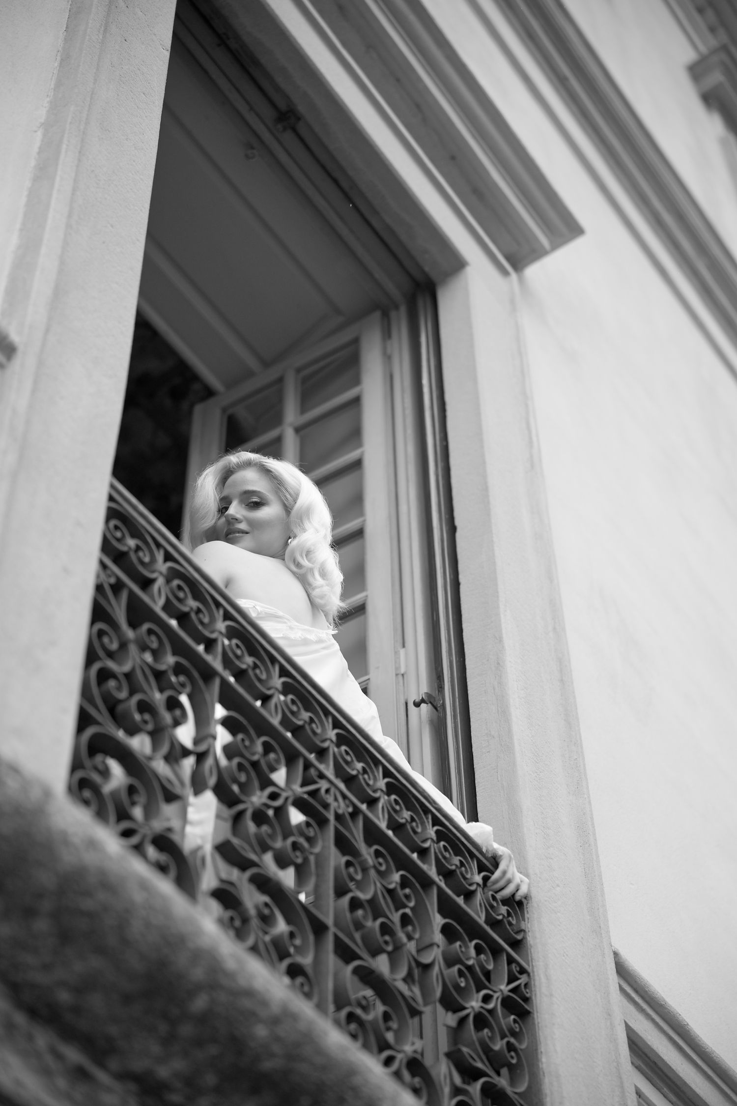 A black and white photograph of a woman standing on an ornate balcony with wrought iron railings.