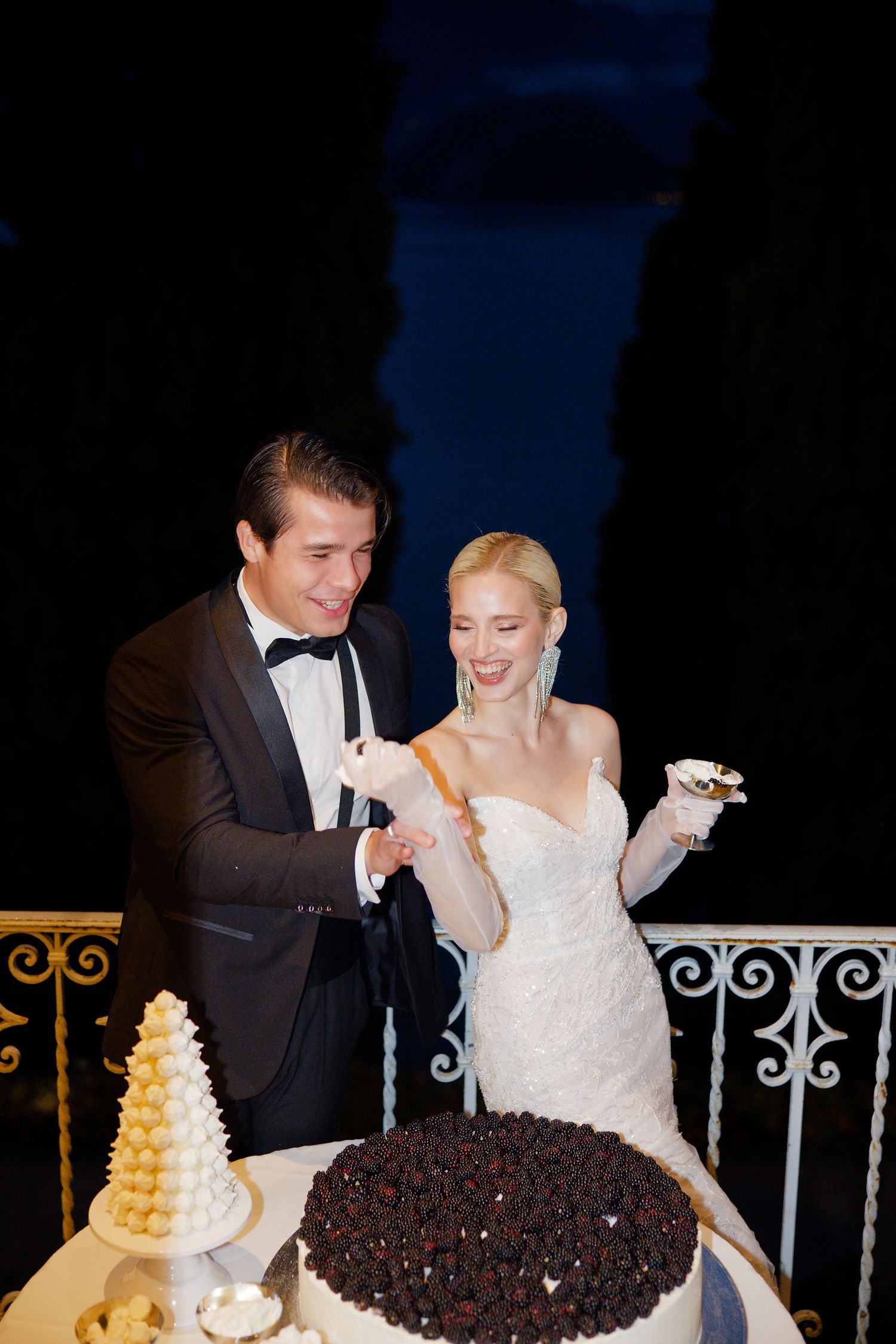 A couple shares their wedding cake together at an elegant outdoor reception with ornate railing details.