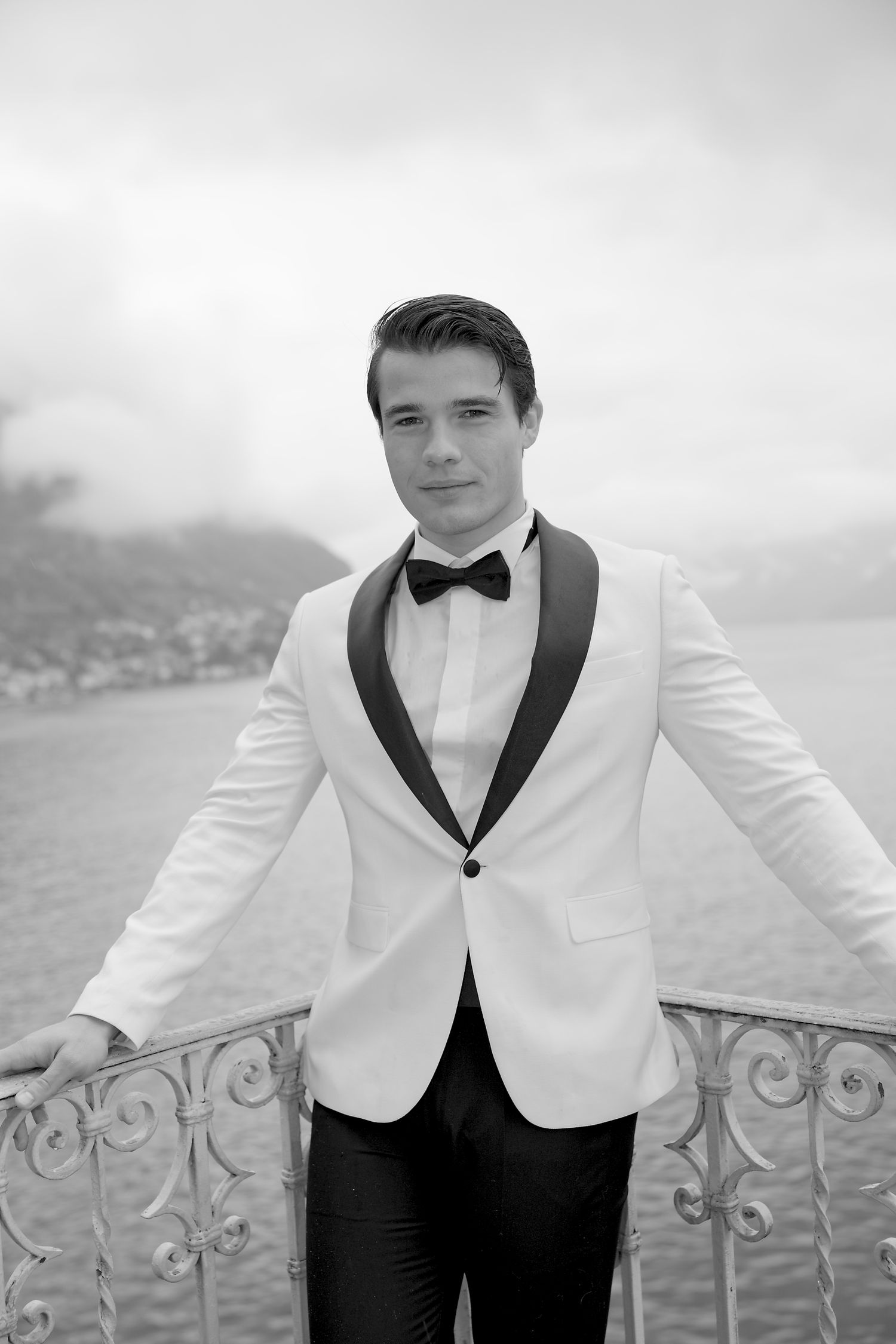 Groom in white dinner jacket poses on balcony overlooking scenic lake view.