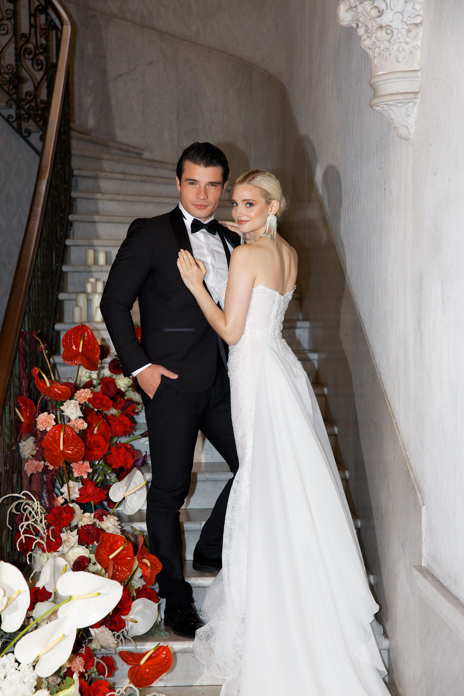 A couple poses on stairs decorated with red and white floral arrangements in an elegant venue.
