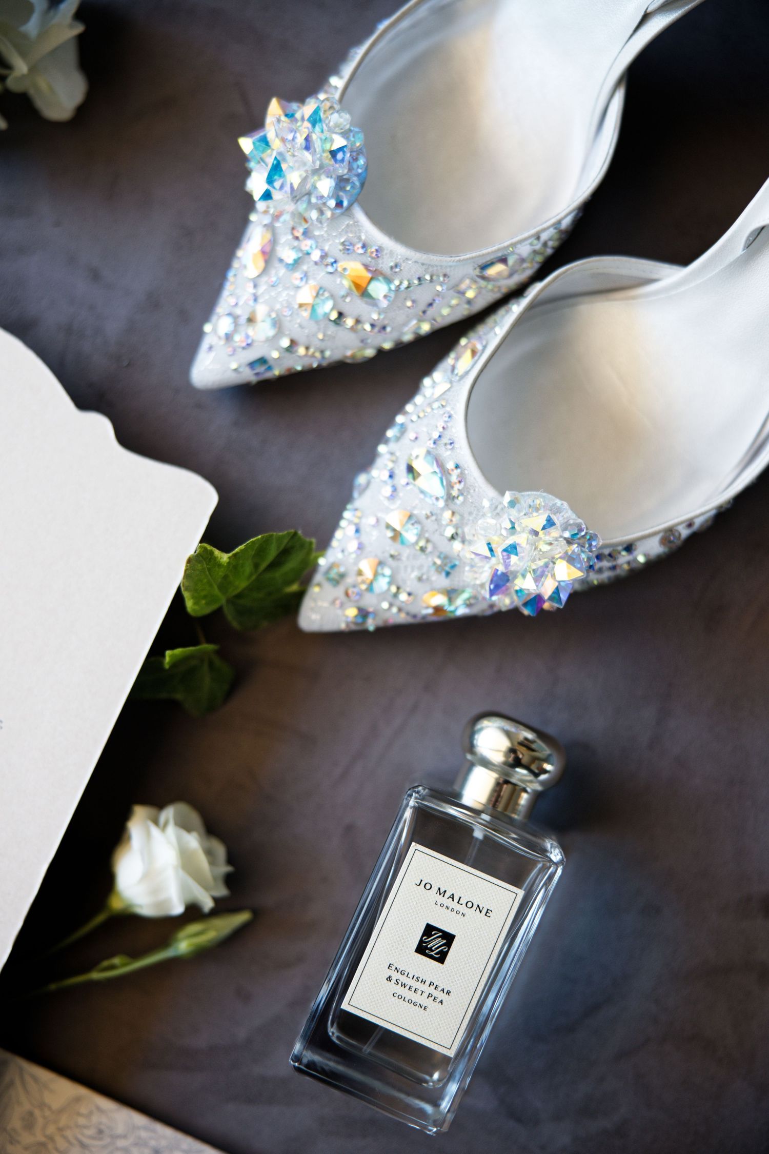 Crystal-encrusted bridal shoes alongside a Jo Malone perfume bottle and white roses on a dark surface.