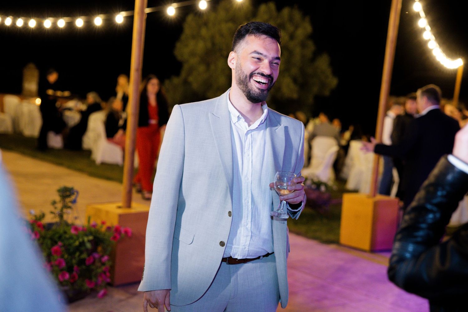 A guest in a light gray suit smiles at an outdoor evening reception with string lights overhead.
