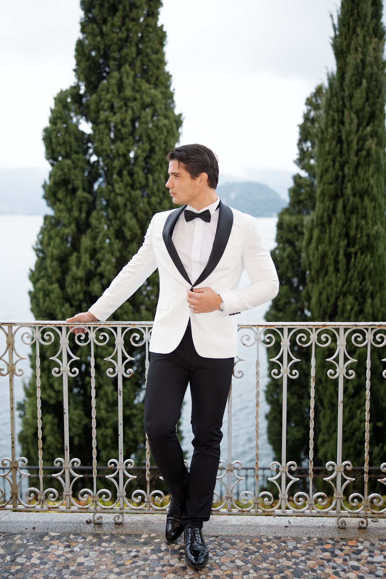 Groom in white tuxedo jacket poses on Mediterranean balcony with cypress trees.