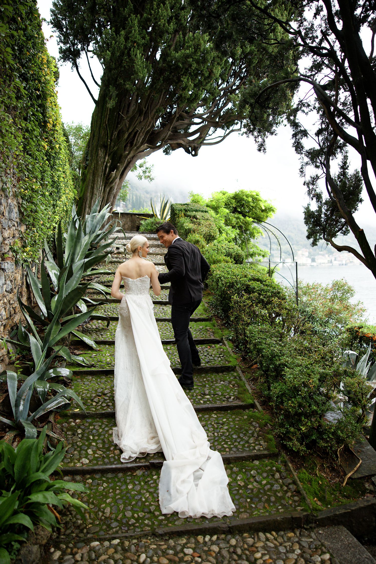 Newlyweds ascend stone garden steps surrounded by lush vegetation and lake views.
