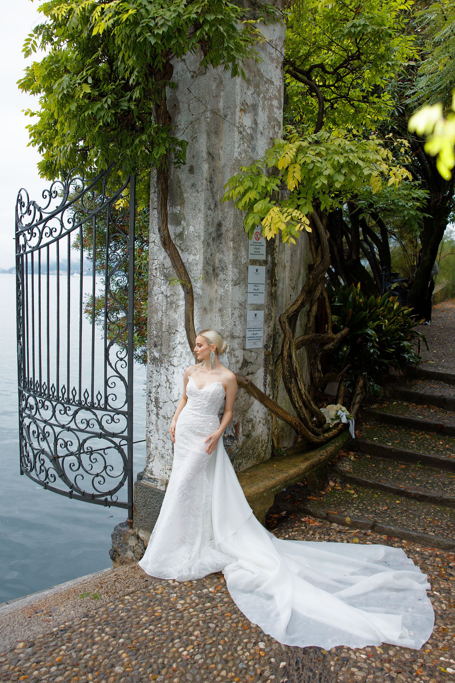Elegant wedding dress with long train photographed against wrought iron gate and stone steps in garden setting.