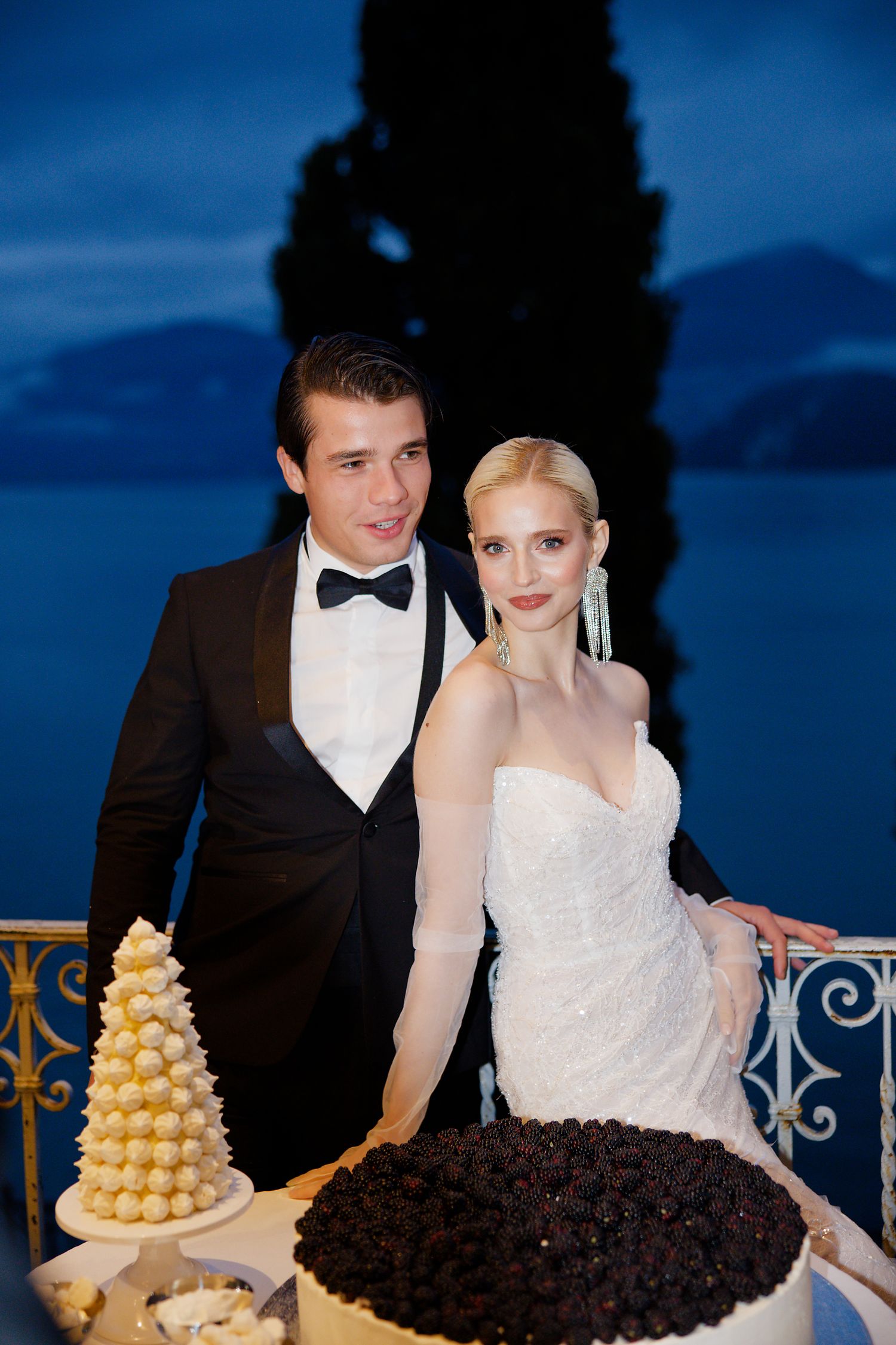 Elegant newlyweds pose beside their wedding cake display with a yellow macaron tower during twilight.