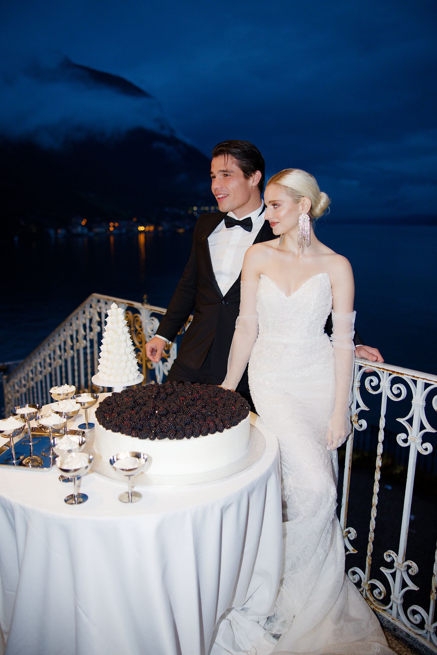 Evening wedding reception on lakeside terrace with two-tiered cake display and scenic mountain views.