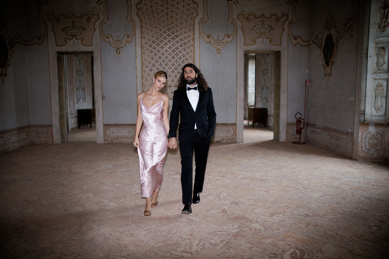Newlywed couple walking together through an ornate ballroom in formal wedding attire.