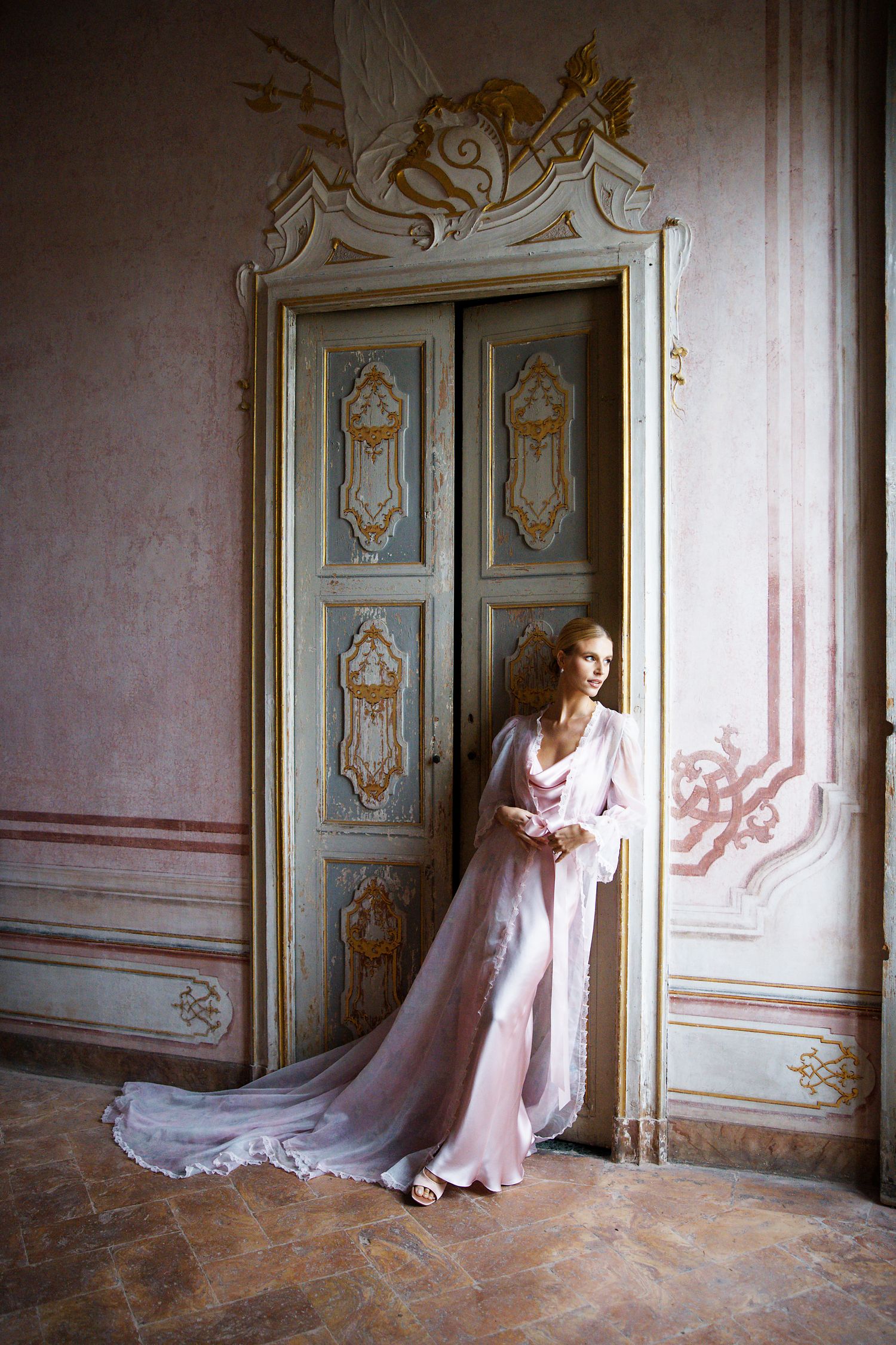 Woman in flowing pink gown stands by ornate vintage door in elegant palace interior.