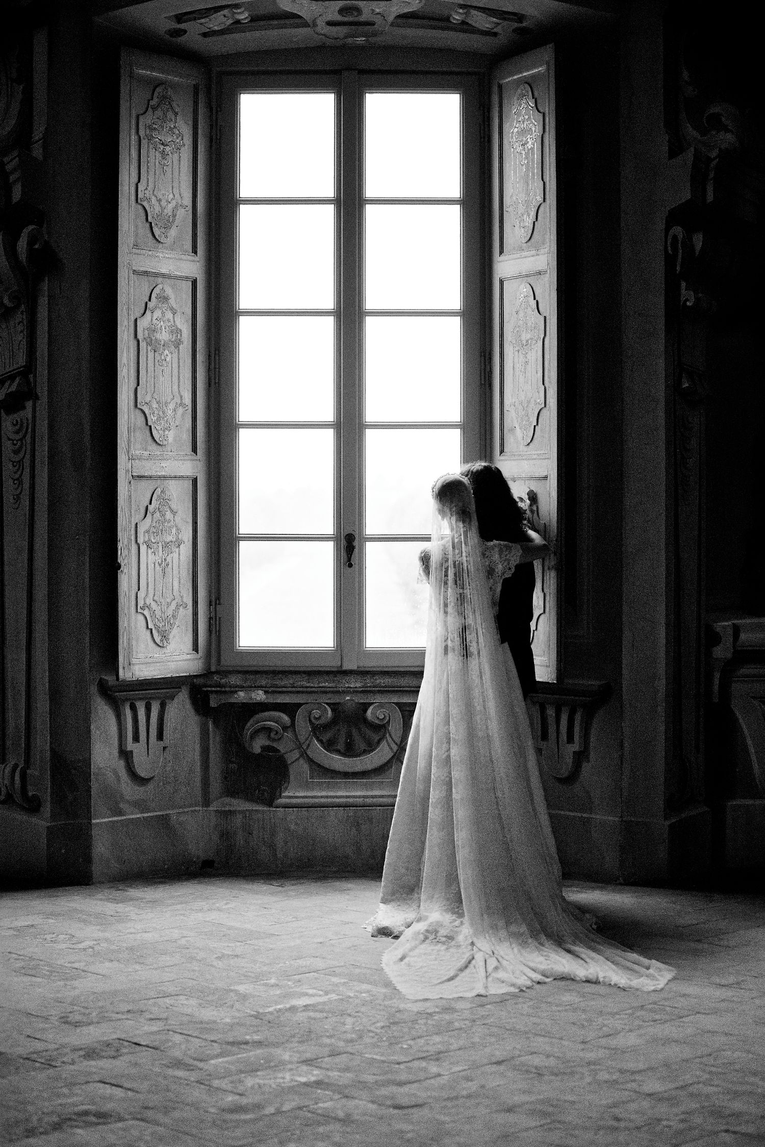 Silhouette of bride in flowing dress and veil gazing through tall palace window.