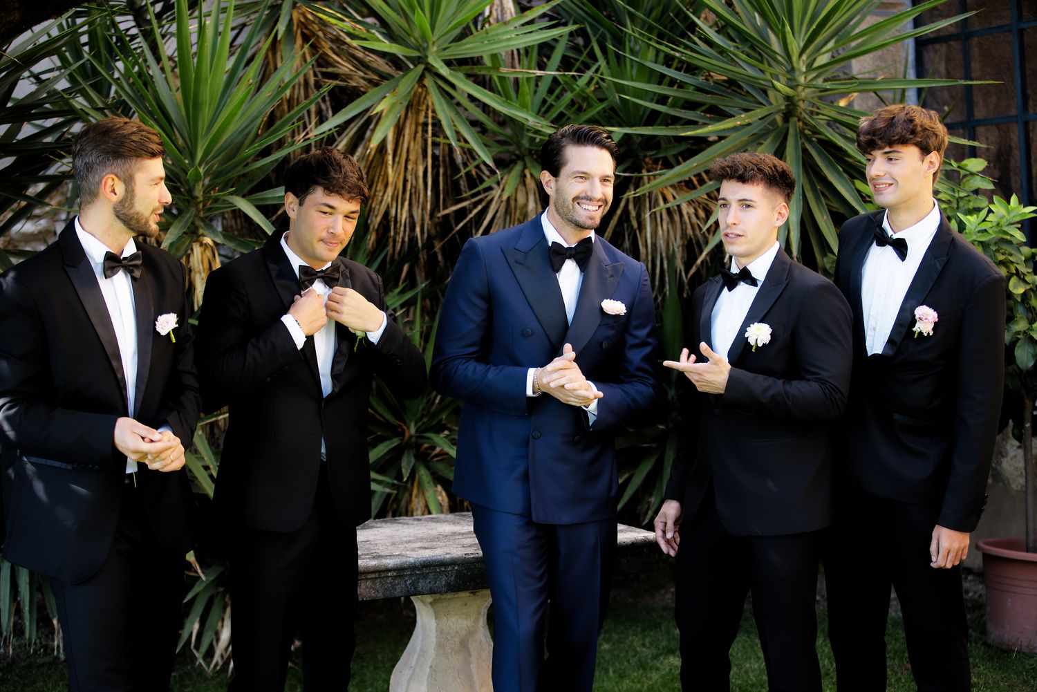 Groomsmen in formal attire share a lighthearted moment together in a garden setting.