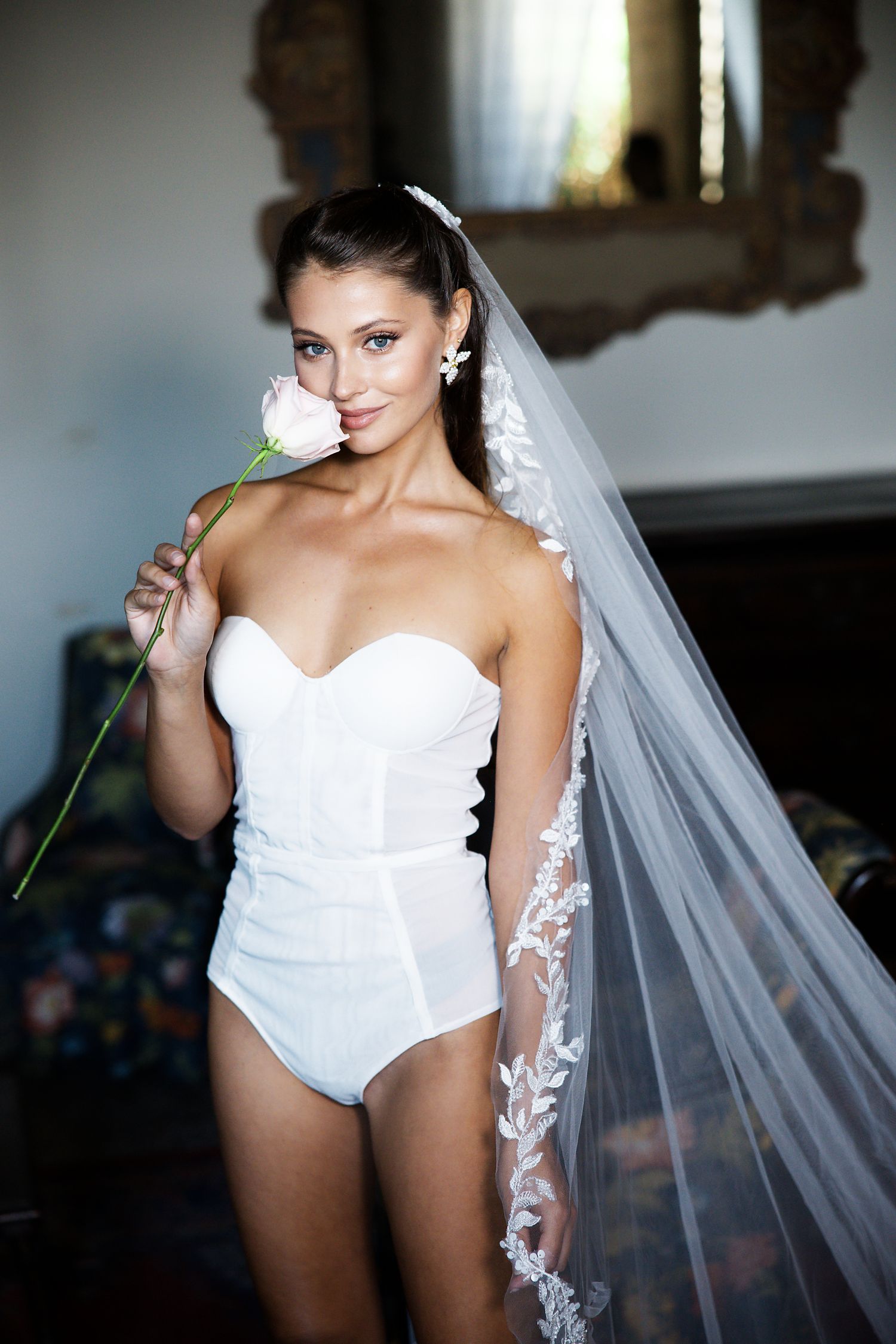 A bride in a white bodysuit holds a single flower while wearing a delicate lace veil.