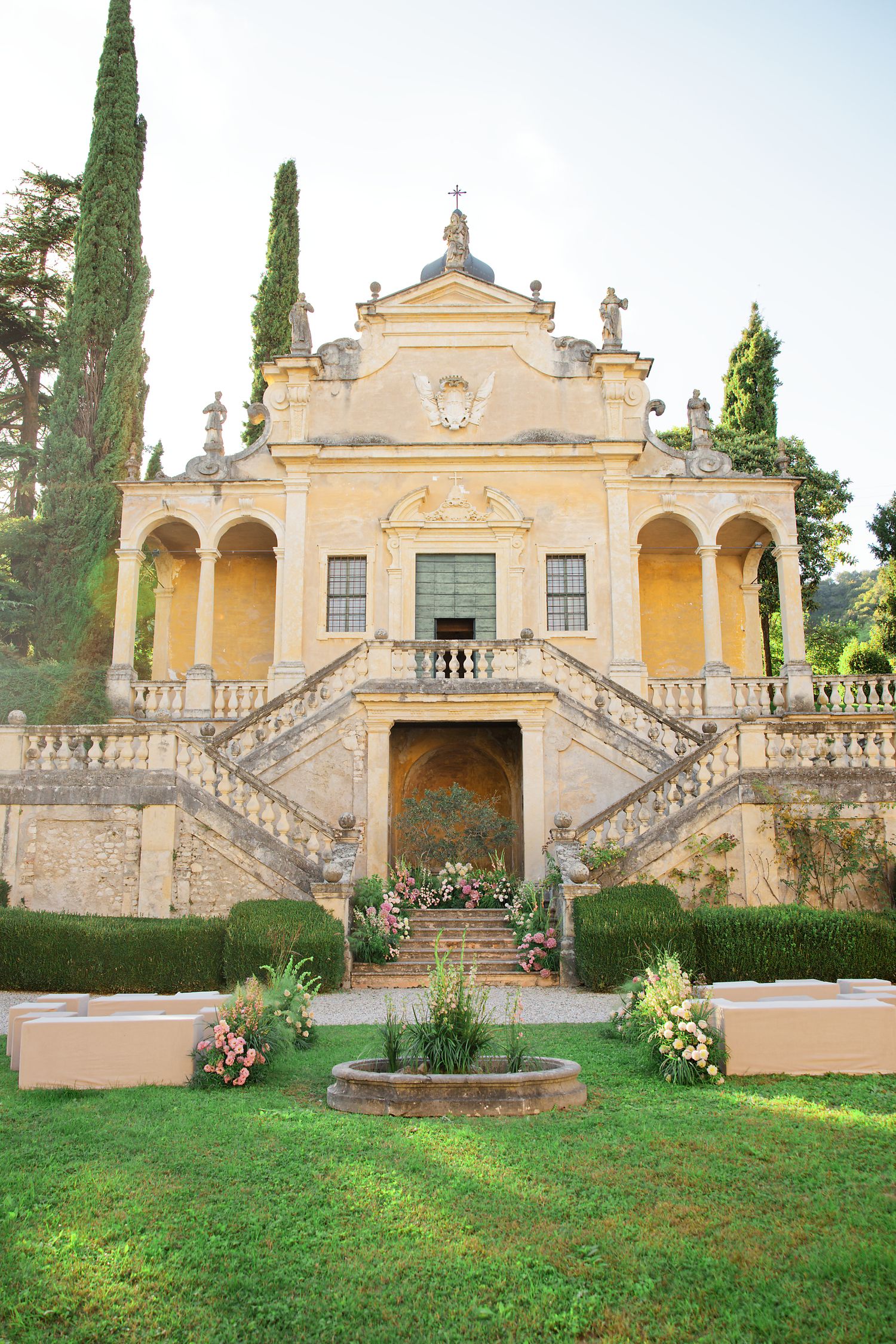 Grand yellow Italian villa with symmetrical staircase and cypress trees in a manicured garden.