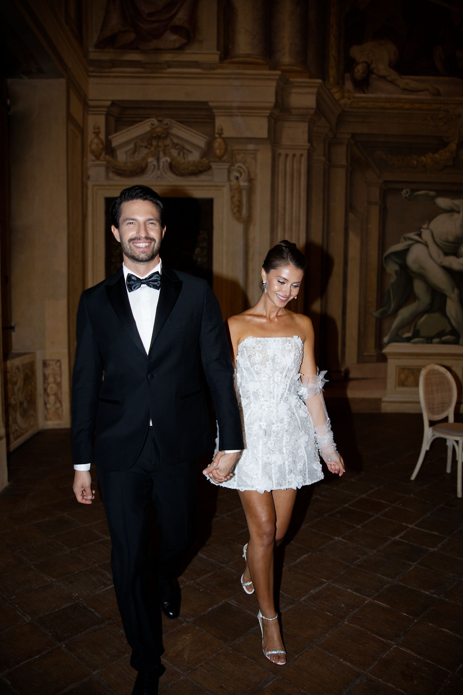 A couple in formal attire walking through an ornate historic venue.