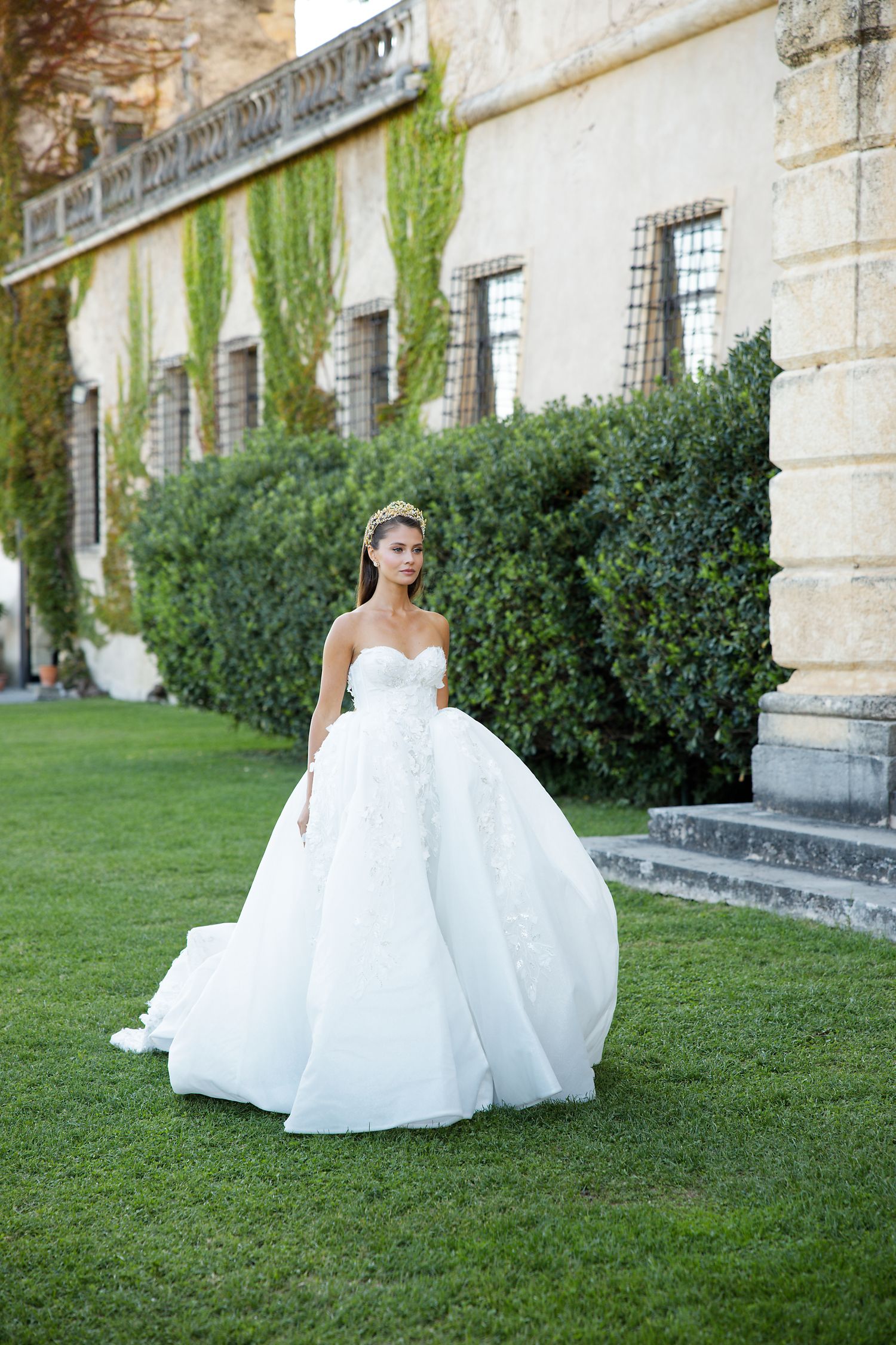 Stunning wedding dress with voluminous ball gown skirt photographed on manicured lawn against historic stone building.