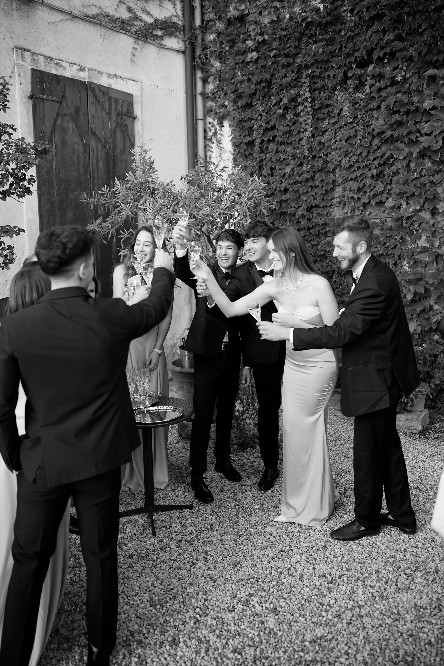 Wedding party toasting with champagne glasses in a black and white photo against an ivy wall.