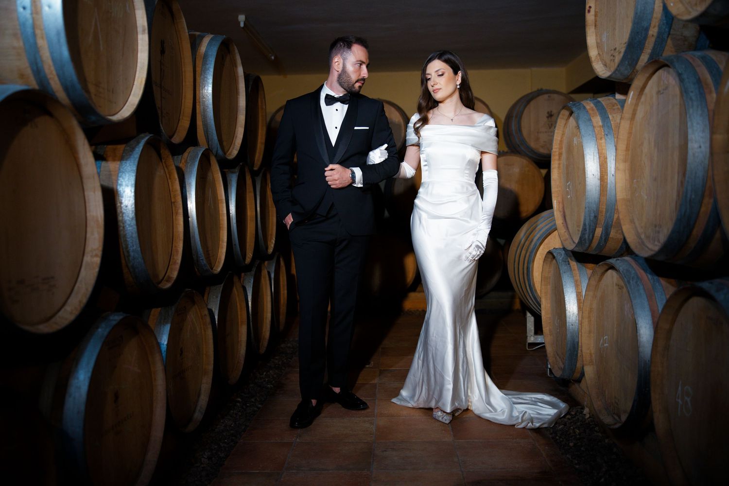 Elegant wedding couple poses in wine cellar surrounded by wooden barrels.