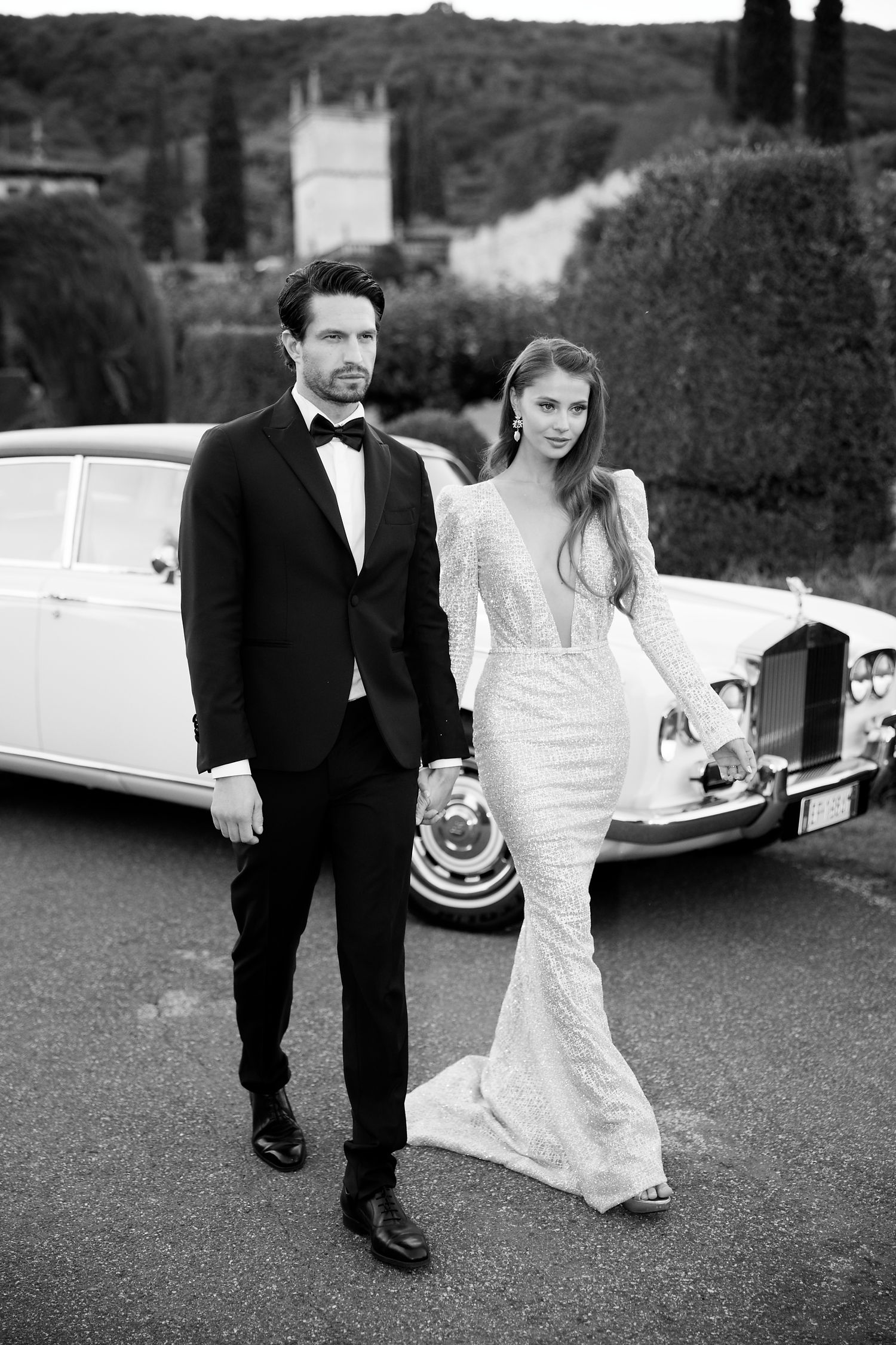 Elegant couple poses in wedding attire next to a classic white luxury car in a black and white photo.