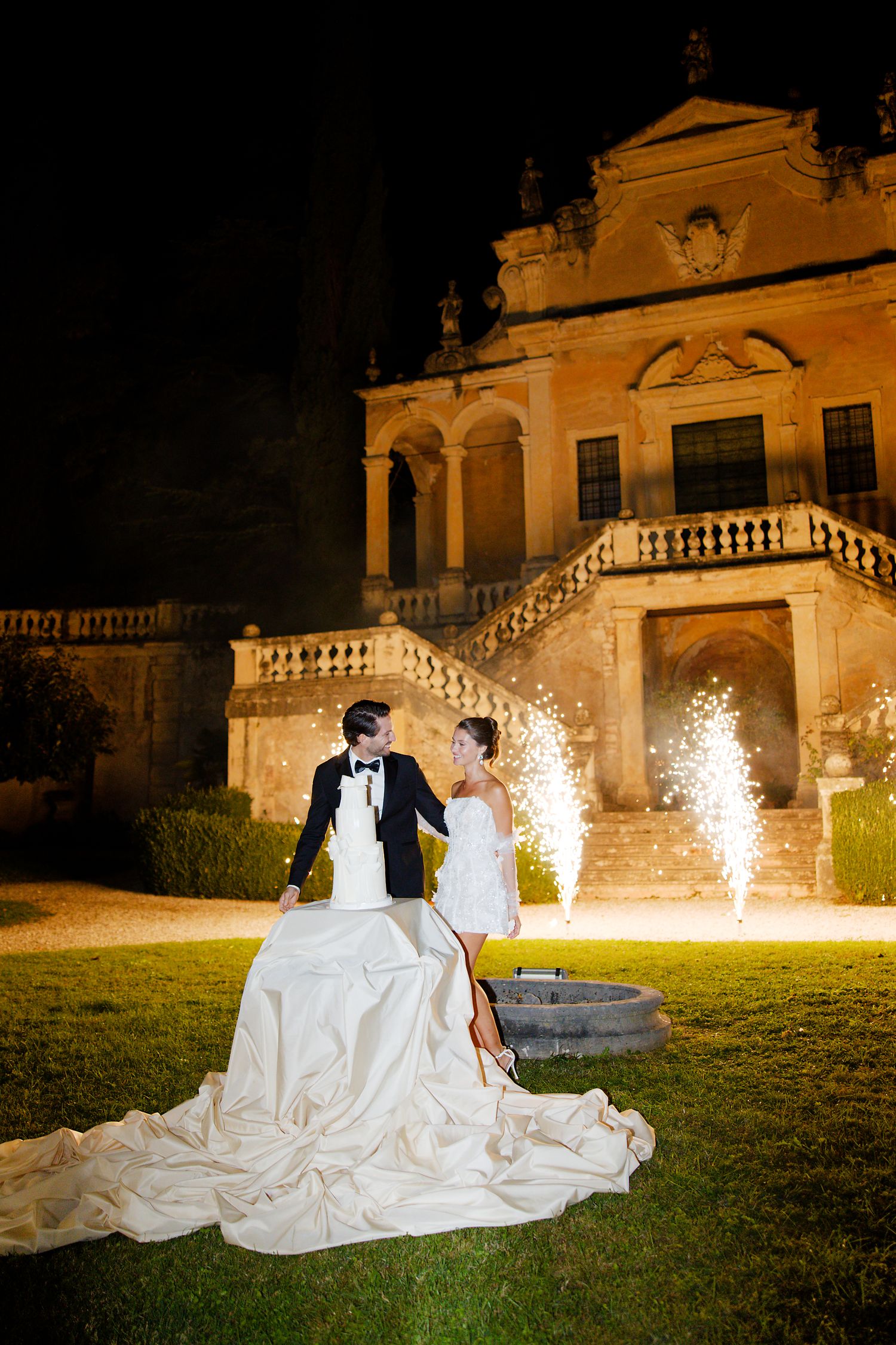 A wedding couple poses in front of an illuminated villa at night with sparkling fireworks in the background.