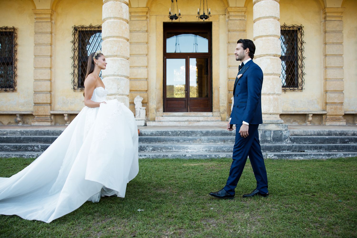 A wedding couple stands on manicured lawn in front of an elegant yellow mansion with classical architecture.