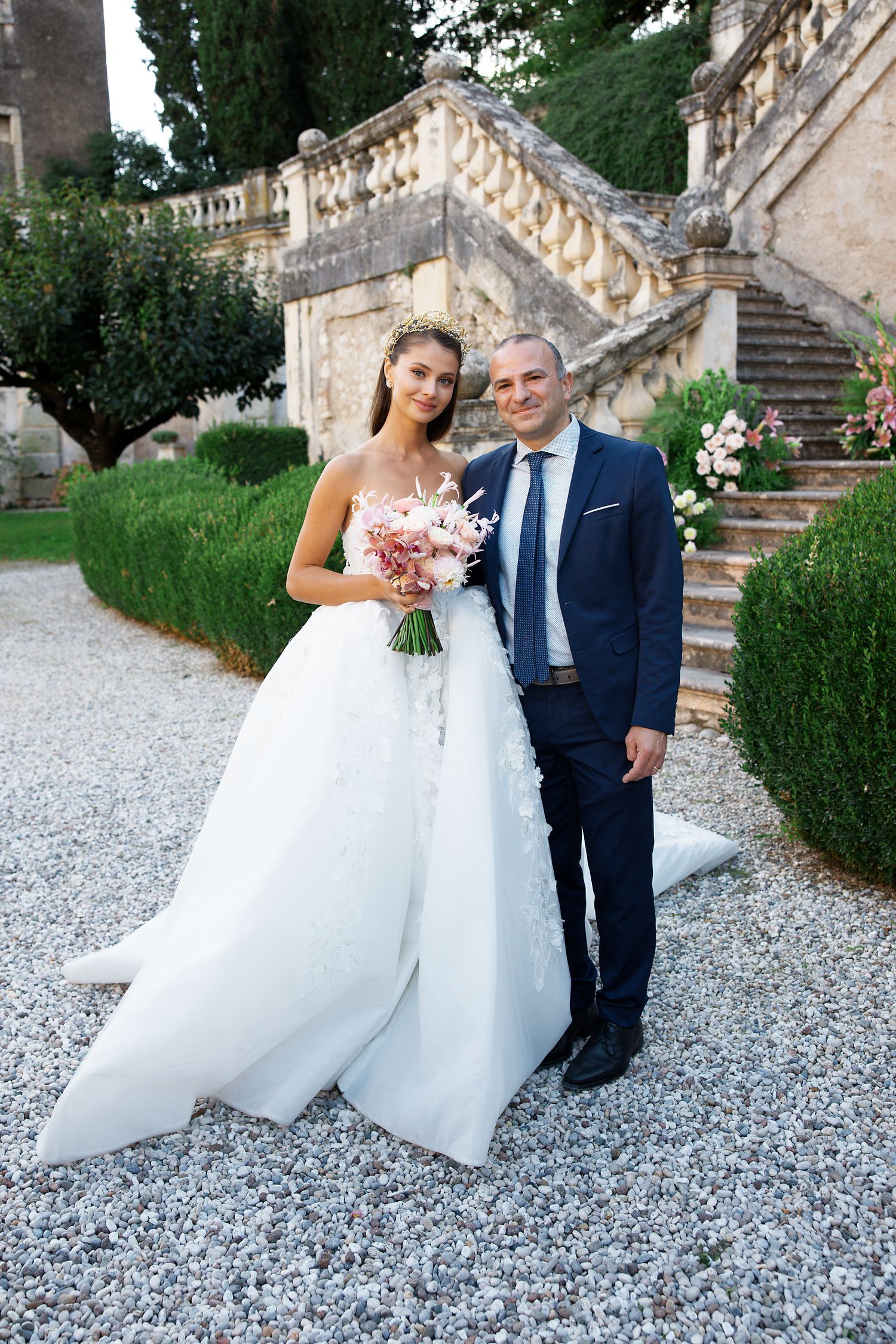 A wedding portrait on gravel path beside ornate stone staircase and manicured gardens.
