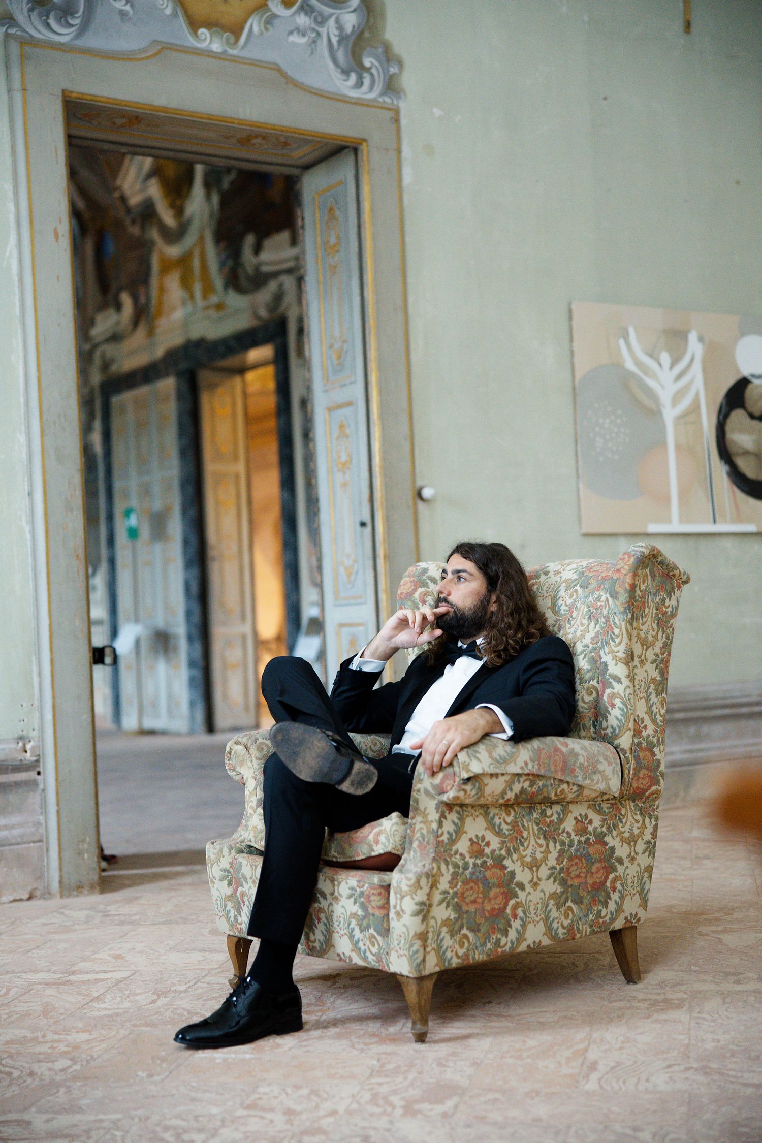 Man in dark suit relaxes thoughtfully in vintage floral armchair inside an elegant room.