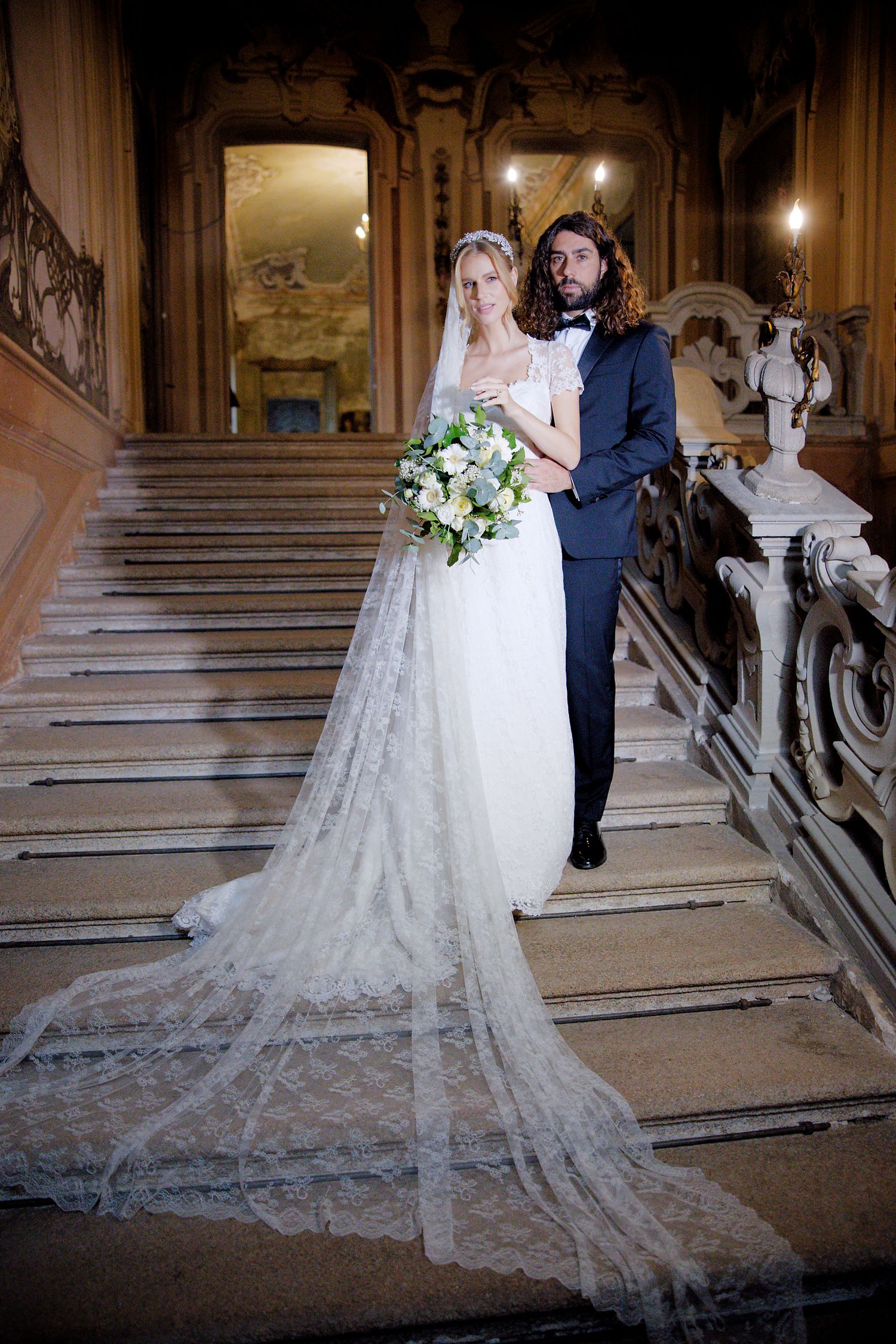 A wedding couple stands on grand marble steps in a palatial setting with decorative baroque architecture.