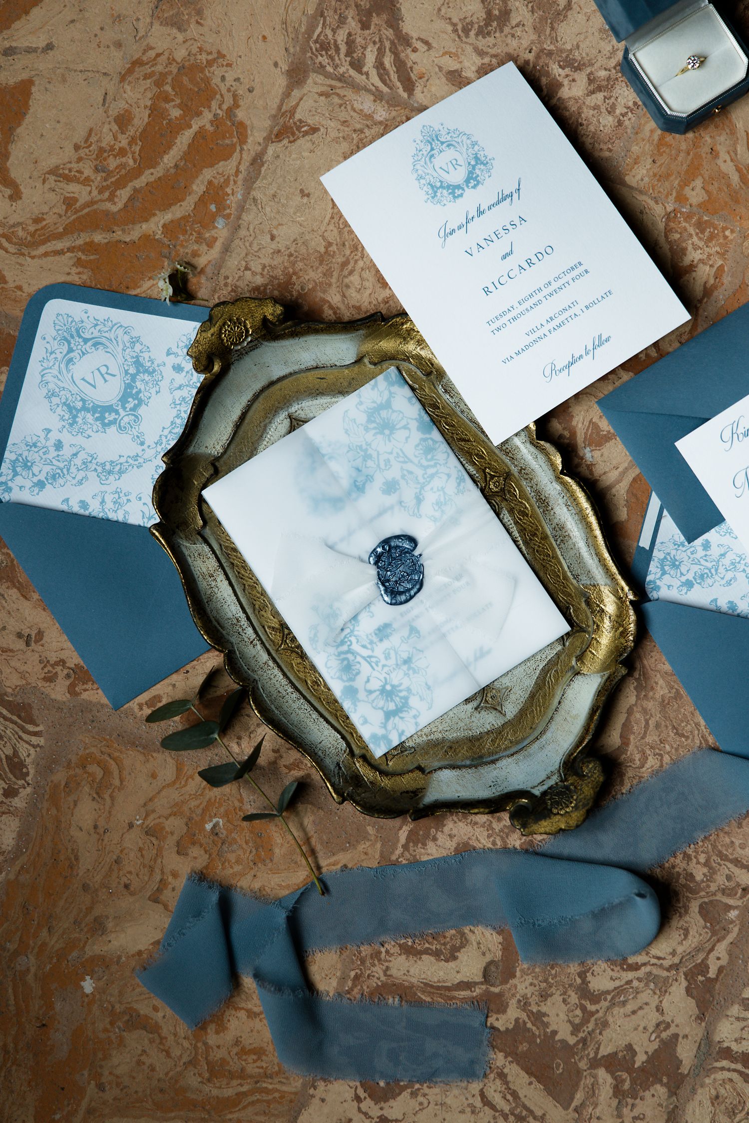 Blue and white wedding invitation with ornate gold frame and wax seal on rustic background.
