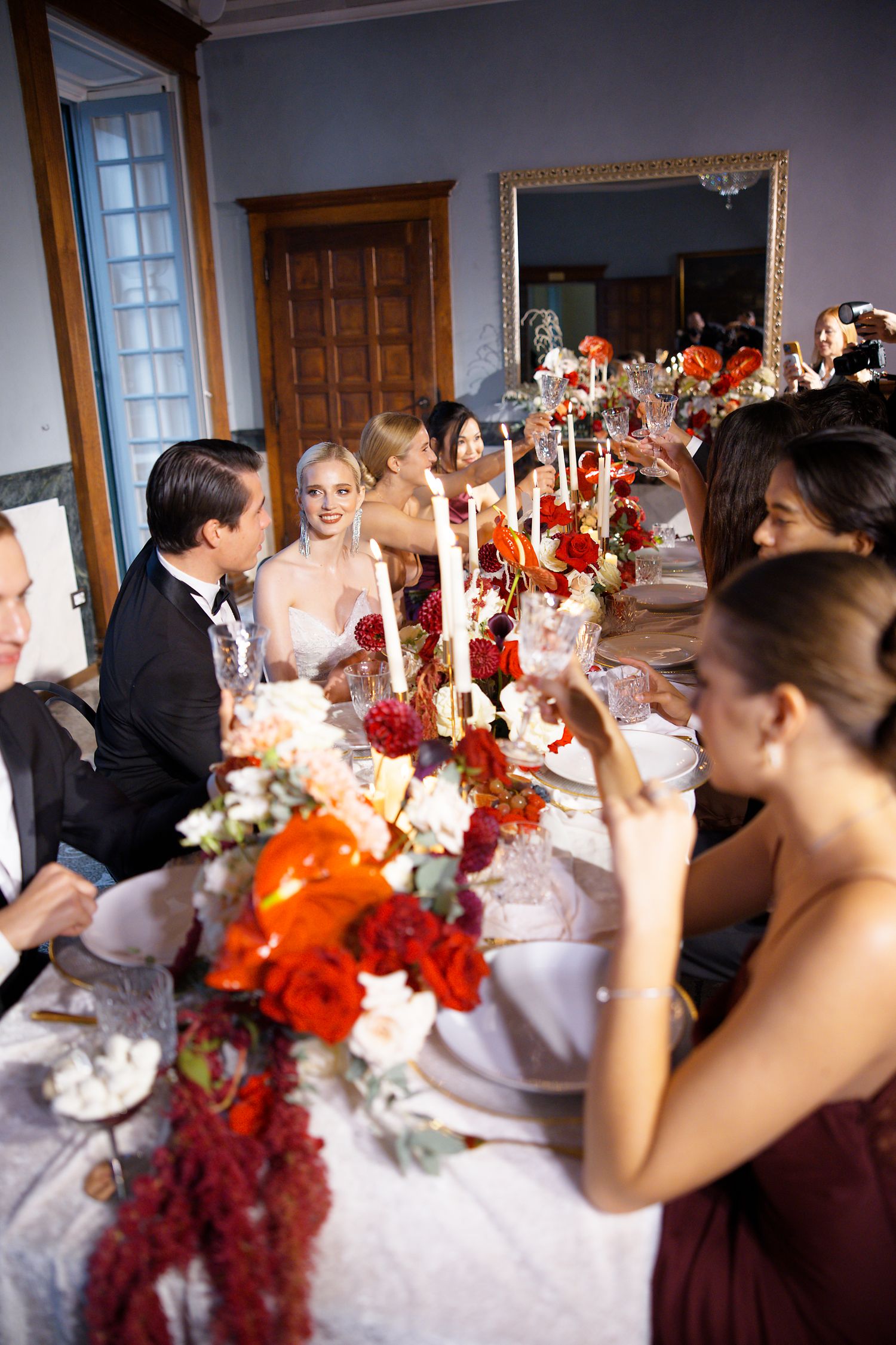 Wedding guests gather around an elegant dinner table decorated with red florals and glowing candles.