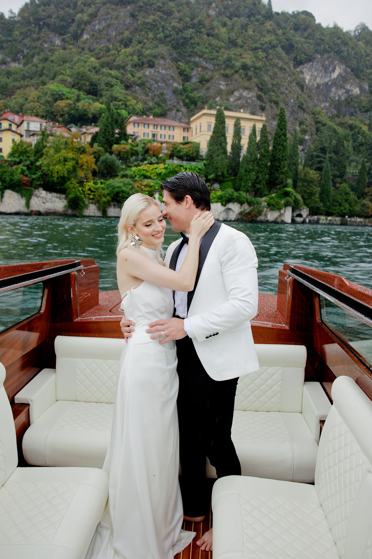 Romantic couple in wedding attire standing on luxury wooden boat on Italian lake.