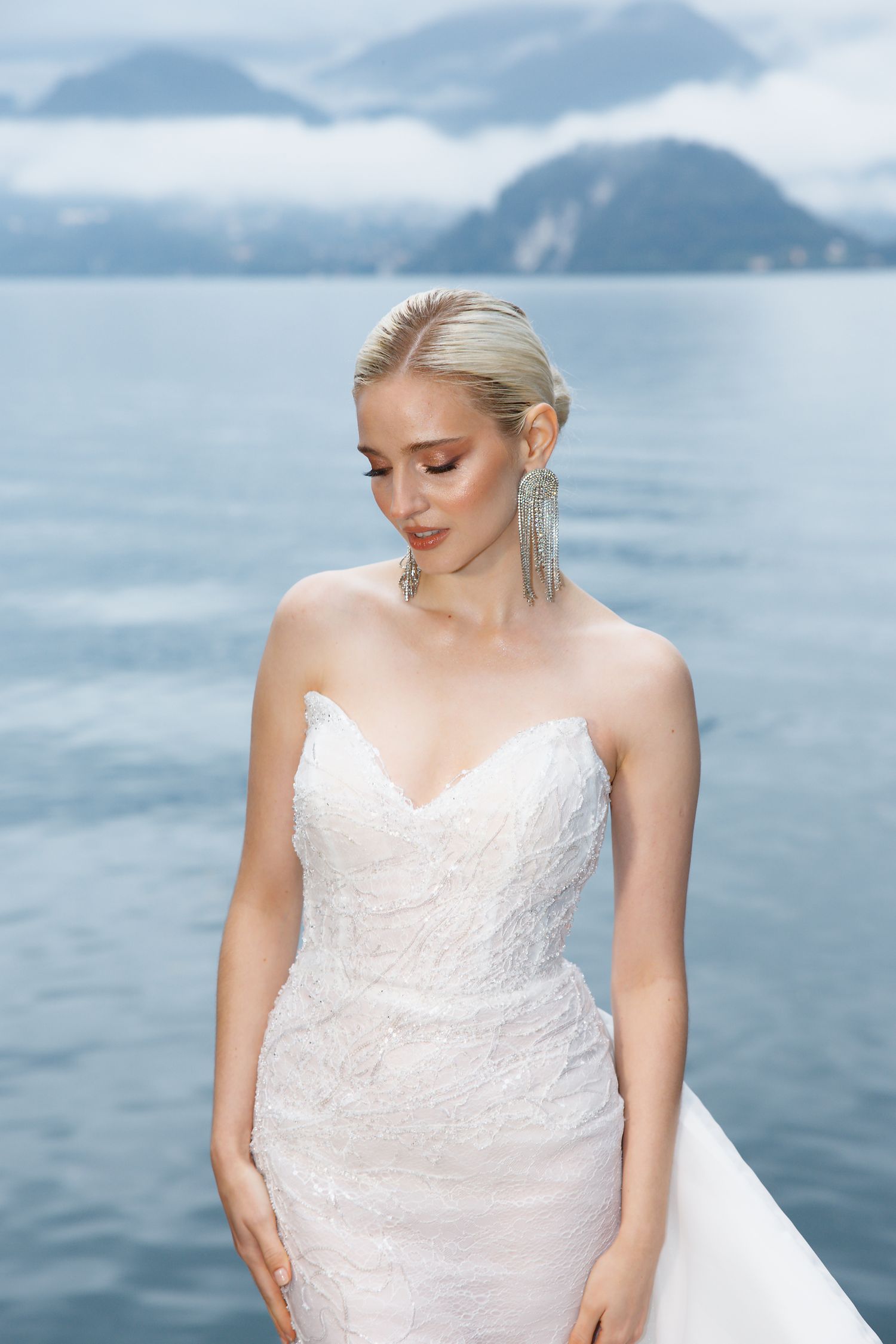 Bride in white lace gown poses against mountain lake backdrop with statement earrings.