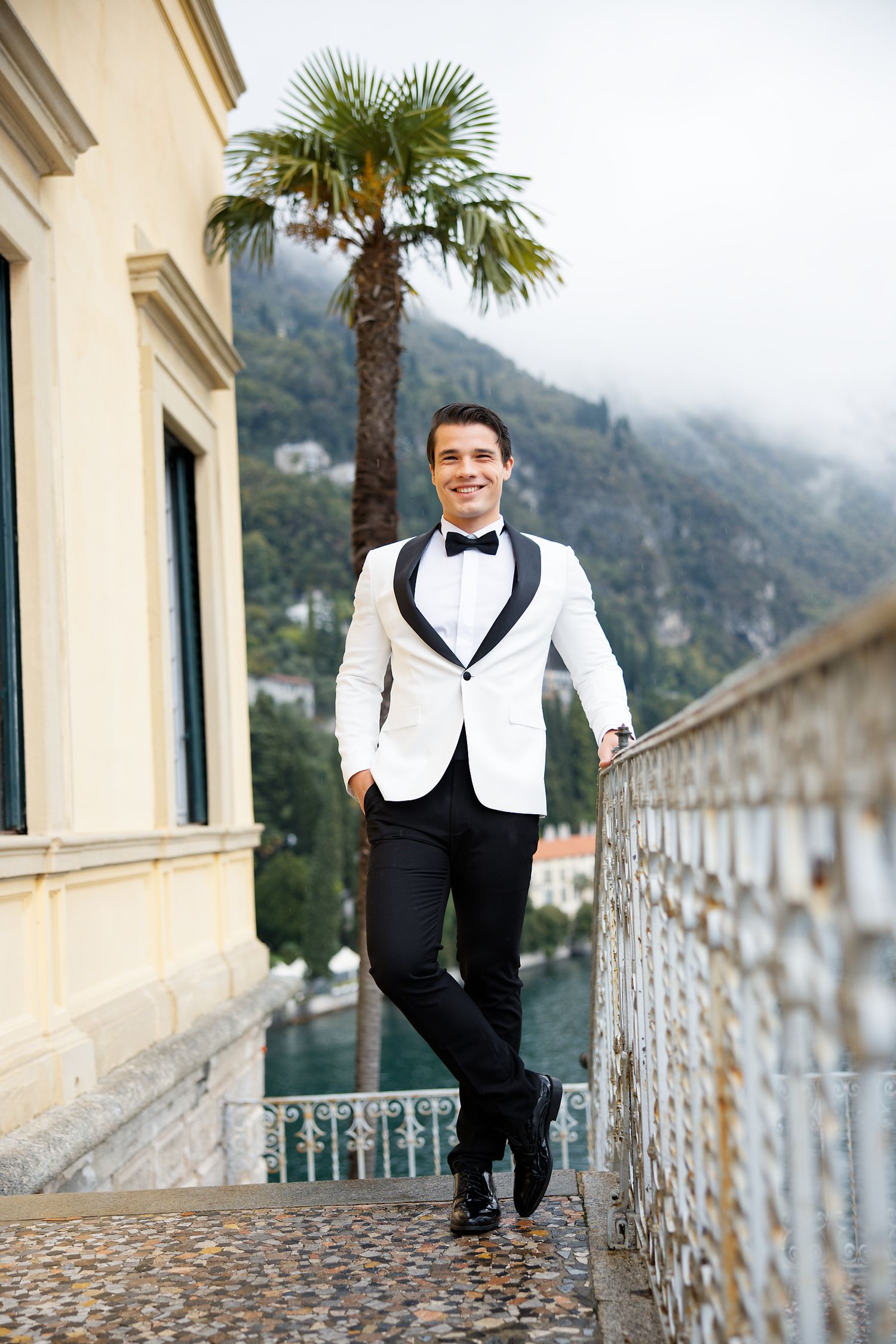 Groom in classic white dinner jacket stands by palm tree on Italian villa terrace.