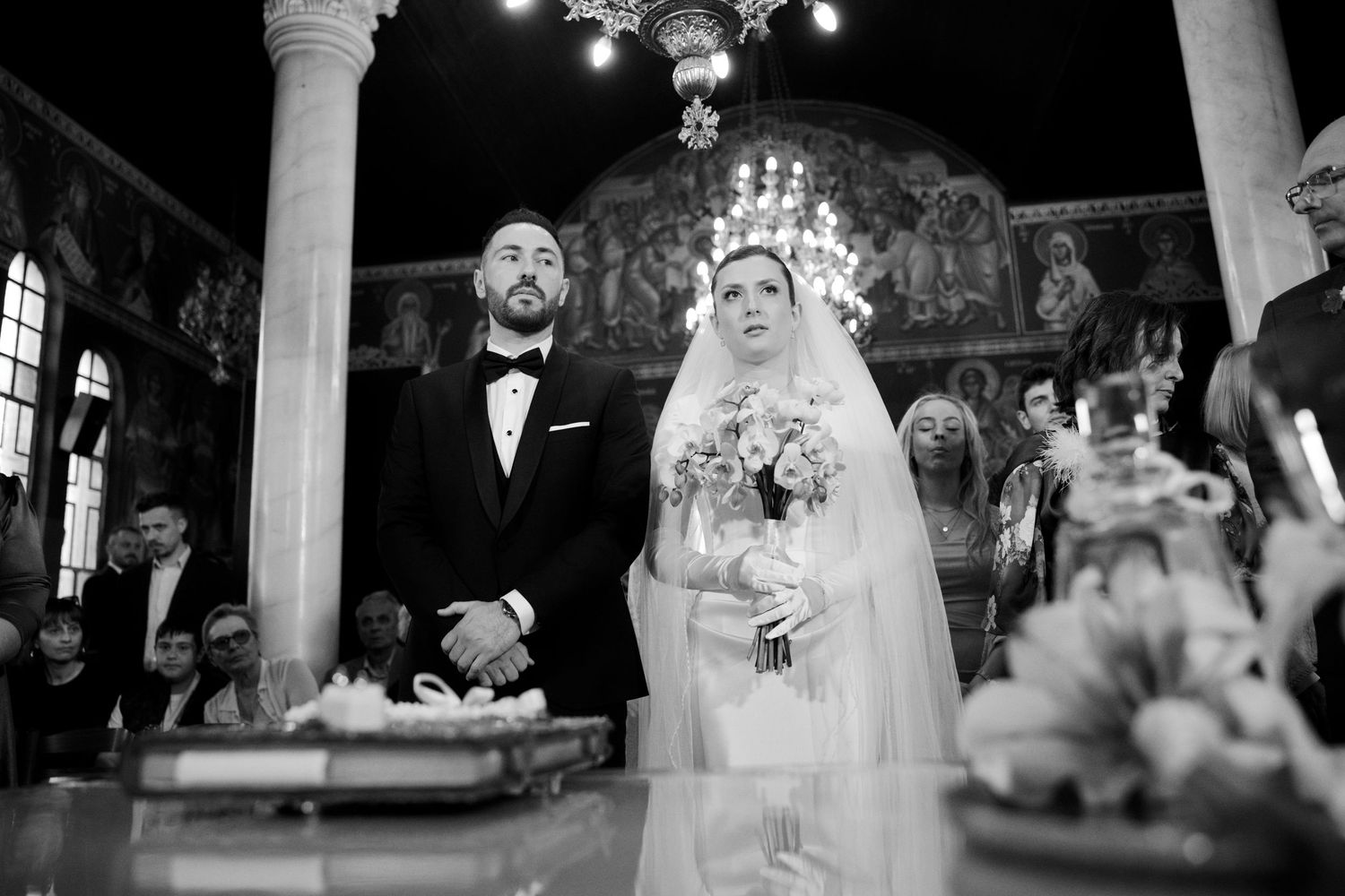 Couple stands at altar during Orthodox wedding ceremony surrounded by ornate decorations and candles.