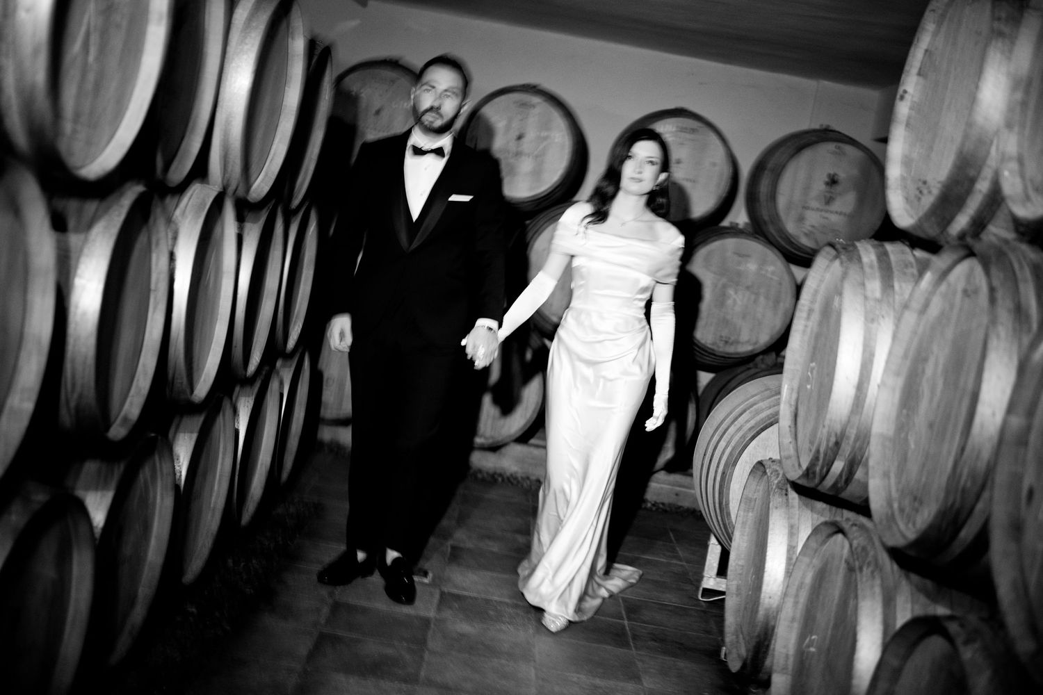 Black and white photo of couple walking through wine cellar with large oak barrels.