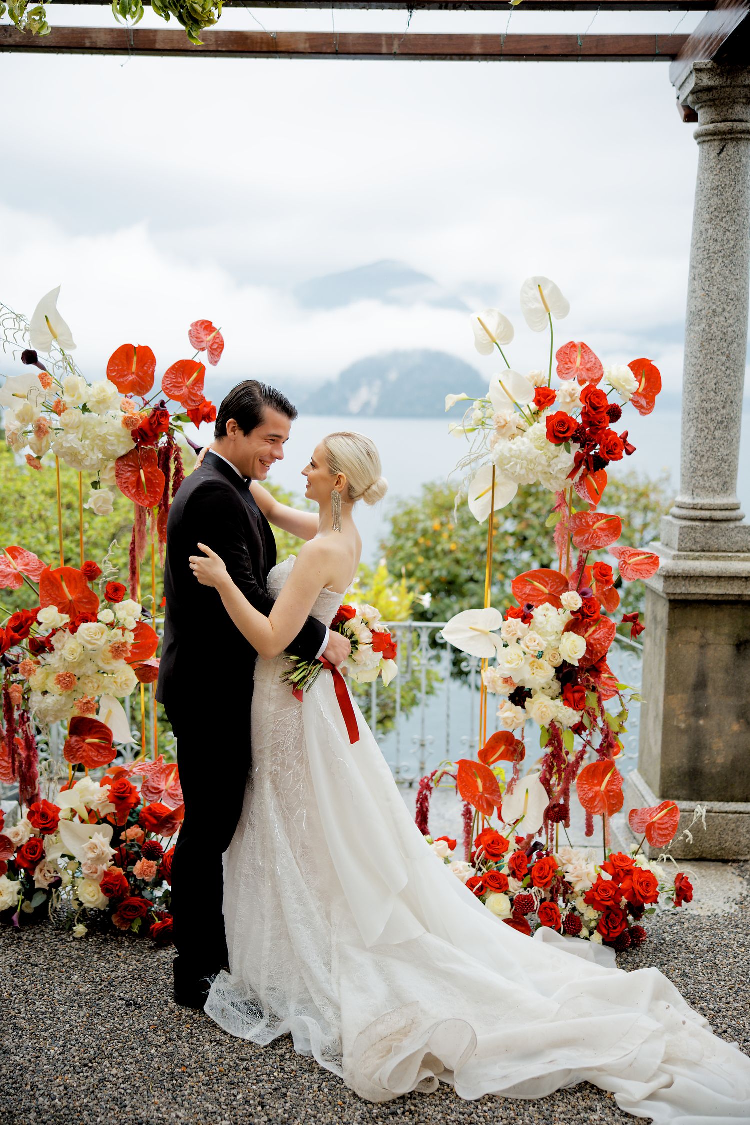 Wedding couple embraces under archway adorned with coral and white floral arrangements overlooking mountain vista.