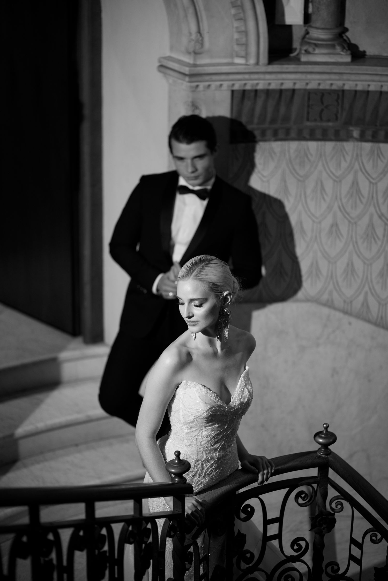 A black and white portrait of a couple in formal attire on an elegant staircase with ornate iron railings.