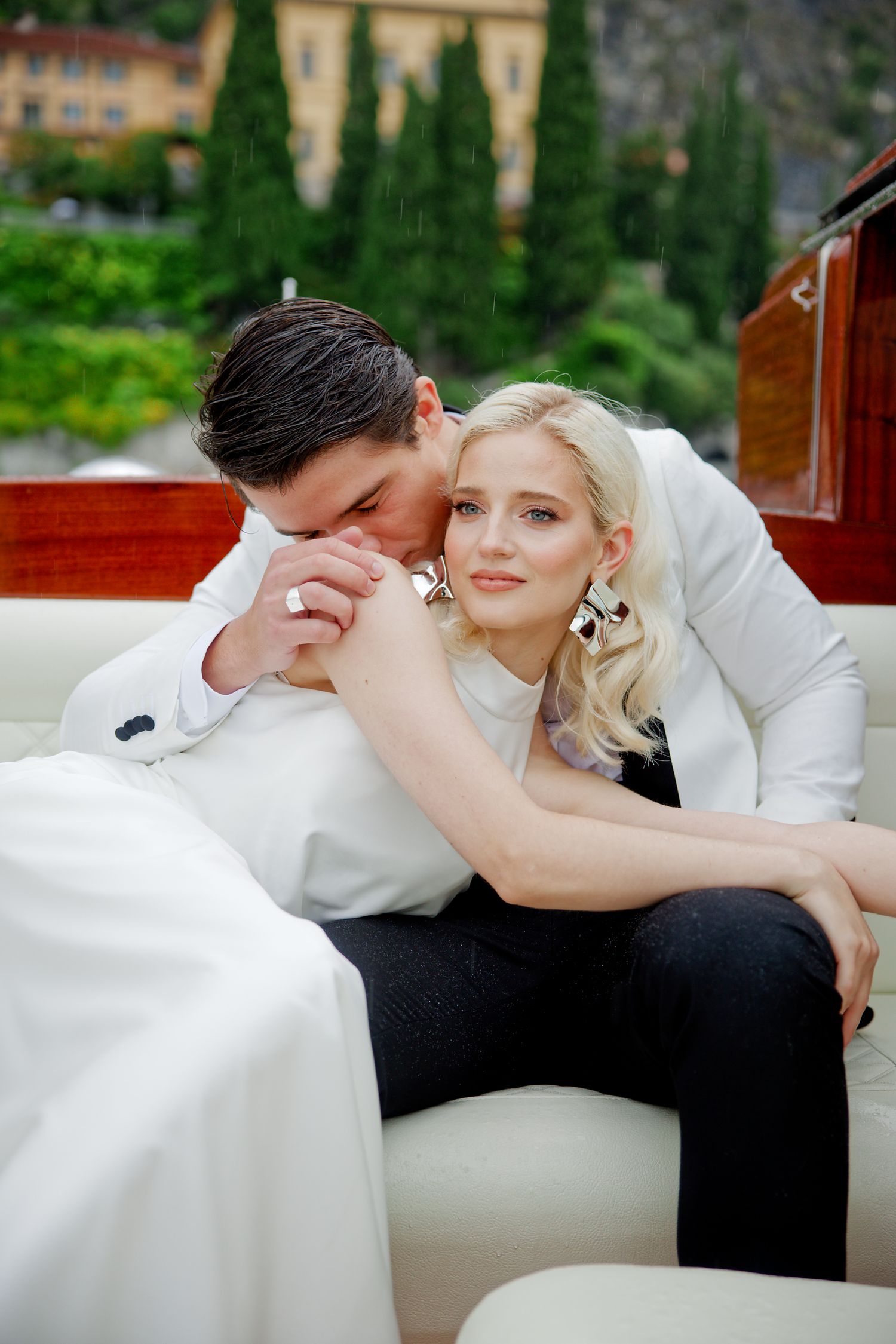 Romantic couple in white attire share an intimate moment on a luxury boat with Italian cypress trees in background.