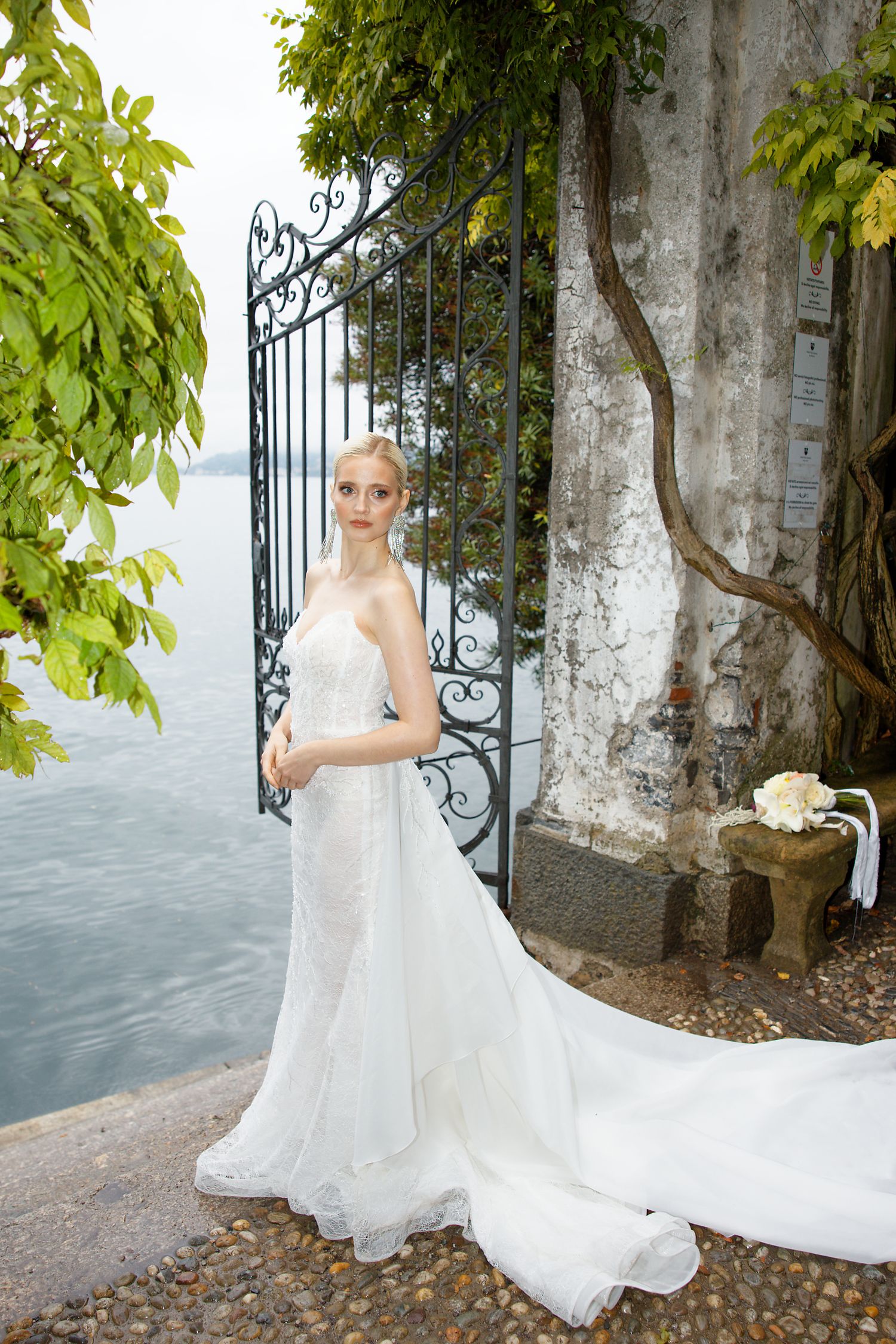 Bride in flowing white dress poses by ornate iron gate with lake view.